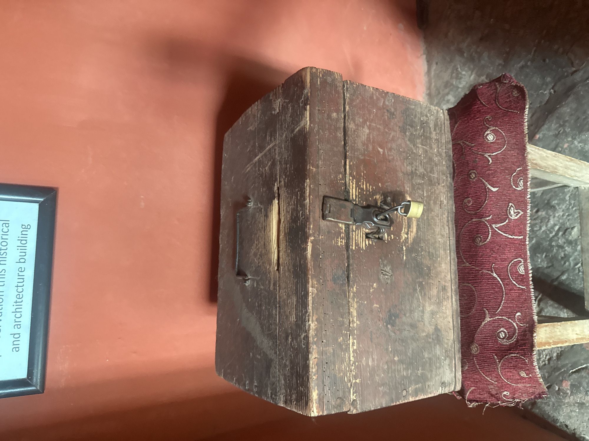 A medium shot depicts a weathered wooden box secured with a padlock, positioned beside a red patterned cushion. The box exhibits signs of age and wear, suggesting it is located in an historical architecture building, possibly within the city of Chynadiiovo, Ukraine. A plaque is visible to the left, though the text is partially obscured, referencing the preservation of "this historical and architecture building."