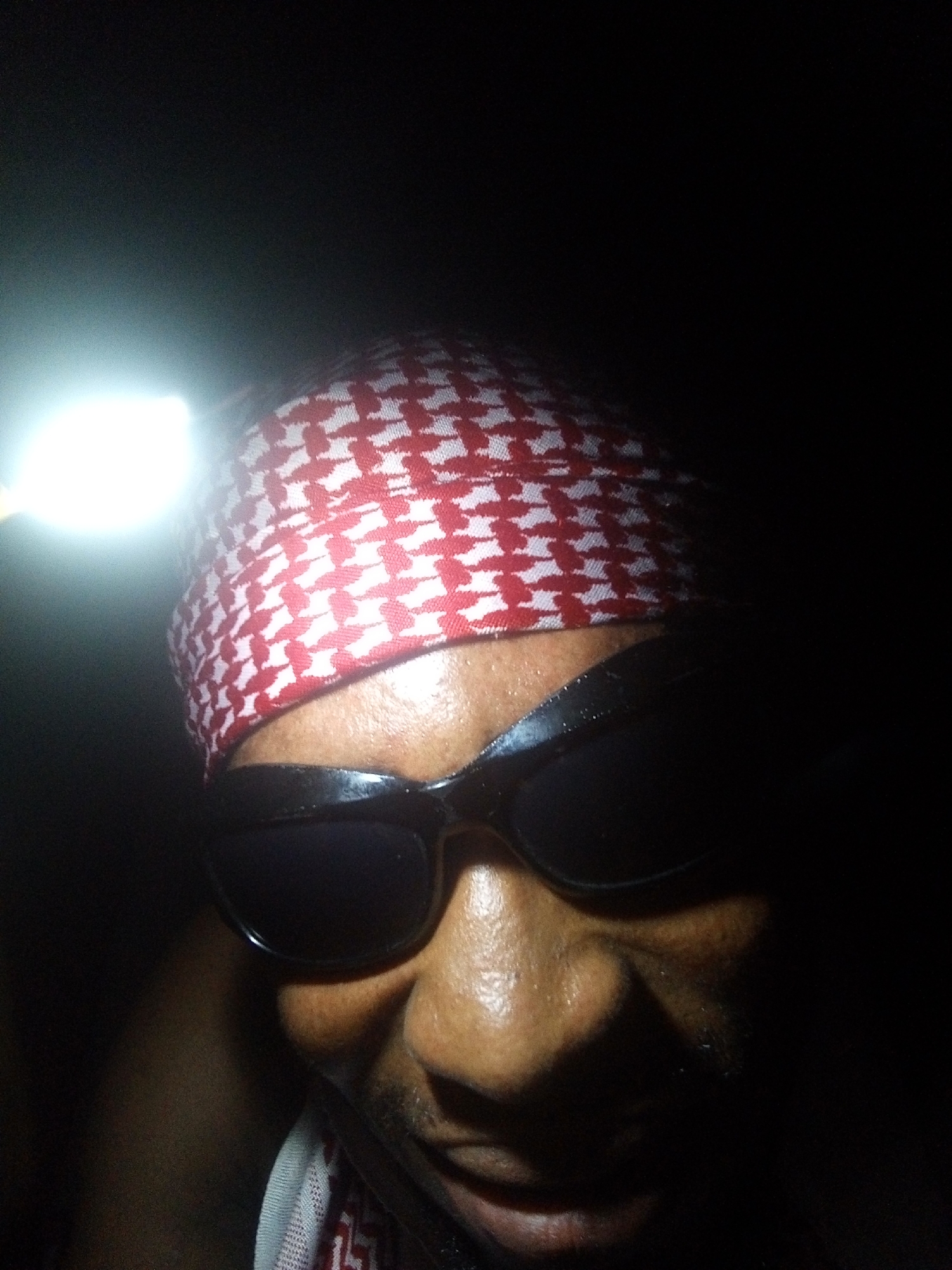 A person wearing a red and white patterned head covering and dark sunglasses is illuminated by a bright light source on their head. The image is taken in low light conditions, with the person's face partially in shadow. No specific location in Lokoja, Nigeria, can be determined from the image.