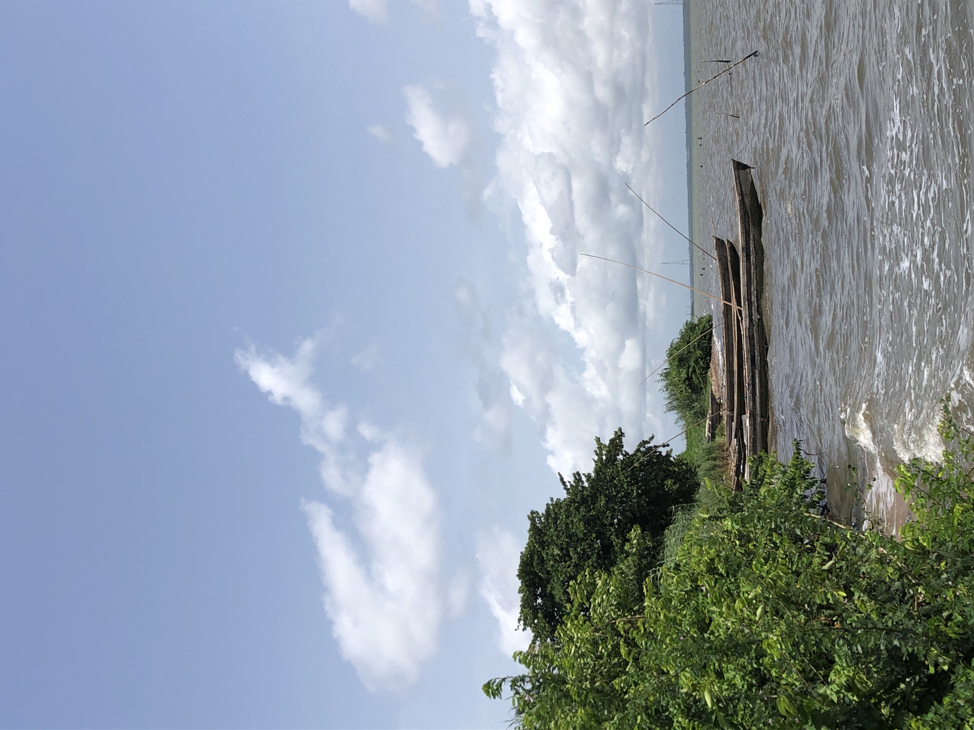 The image depicts a body of water, possibly a lake or river, with a cloudy sky overhead. Along the shoreline, there are two or three narrow boats resting near some green vegetation, including trees and bushes. There are also thin poles or reeds sticking out of the water near the boats. The water appears slightly choppy, with small waves visible near the shore. The location is identified as Unknown, Unknown.