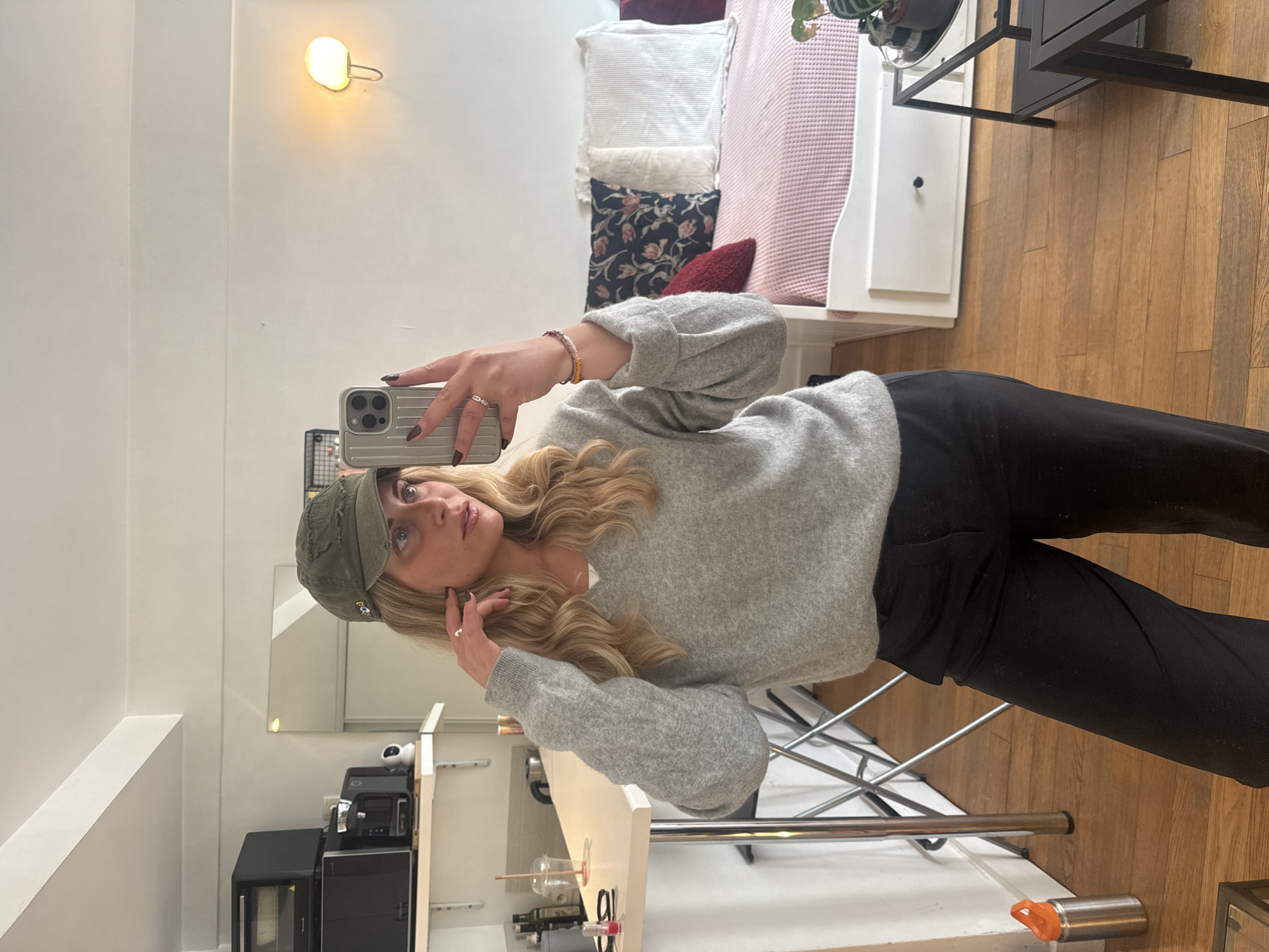 A young woman is taking a selfie in an apartment. She wears a green baseball cap, a light gray sweater, and black pants. The apartment has white walls, a wooden floor, a bed with multiple pillows, and a small kitchen area with a microwave and other appliances. The location could be Paris, France.