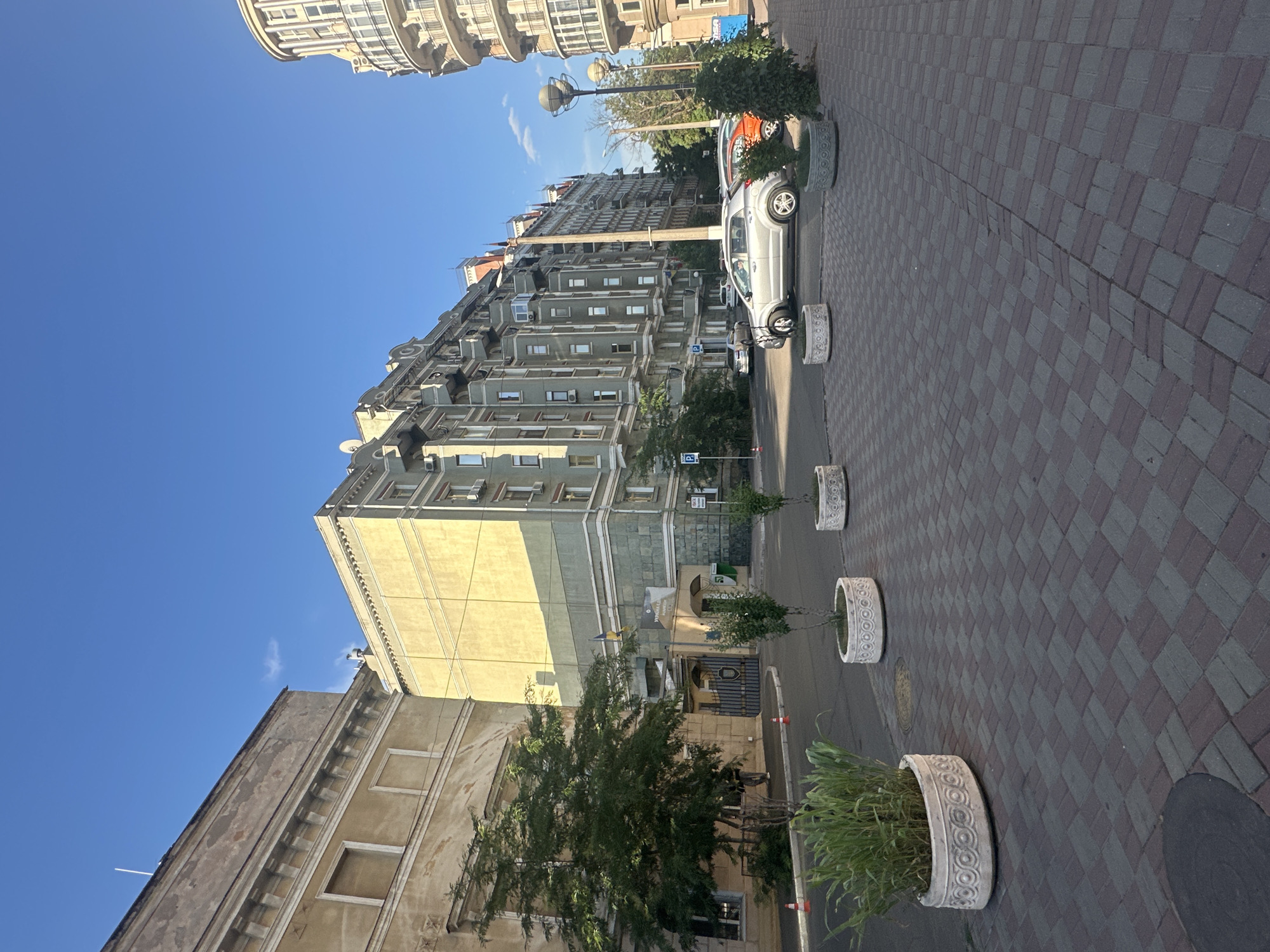 This is a vertical, eye-level shot of a street scene in Odesa, Ukraine, under a clear blue sky, suggesting it's daytime with good weather. The foreground shows a patterned pedestrian walkway made of dark grey and red rectangular paving stones. Along the walkway, three cylindrical planters with decorative patterns are visible. The nearest planter contains green grasses and foliage.
Beyond the walkway is a street with parked cars. A silver car is prominently featured, and behind it, part of a red car can be seen. The street is lined with trees and several buildings. The most prominent building is a multi-story apartment complex with a light green facade, featuring numerous windows and balconies. To the left of this building, a portion of an older, light yellow building with ornate architectural details is visible. Further in the background, another tall building with a more modern appearance can be seen.
Notable details include lampposts along the street, an air conditioning unit visible on one of the buildings, and traffic cones in the distance. There are no people clearly visible in the image. The overall impression is of a sunny day in an urban environment, with a mix of older and more recent architecture. Ukrainian flags are visible on a pole attached to one of the buildings, confirming the location.
