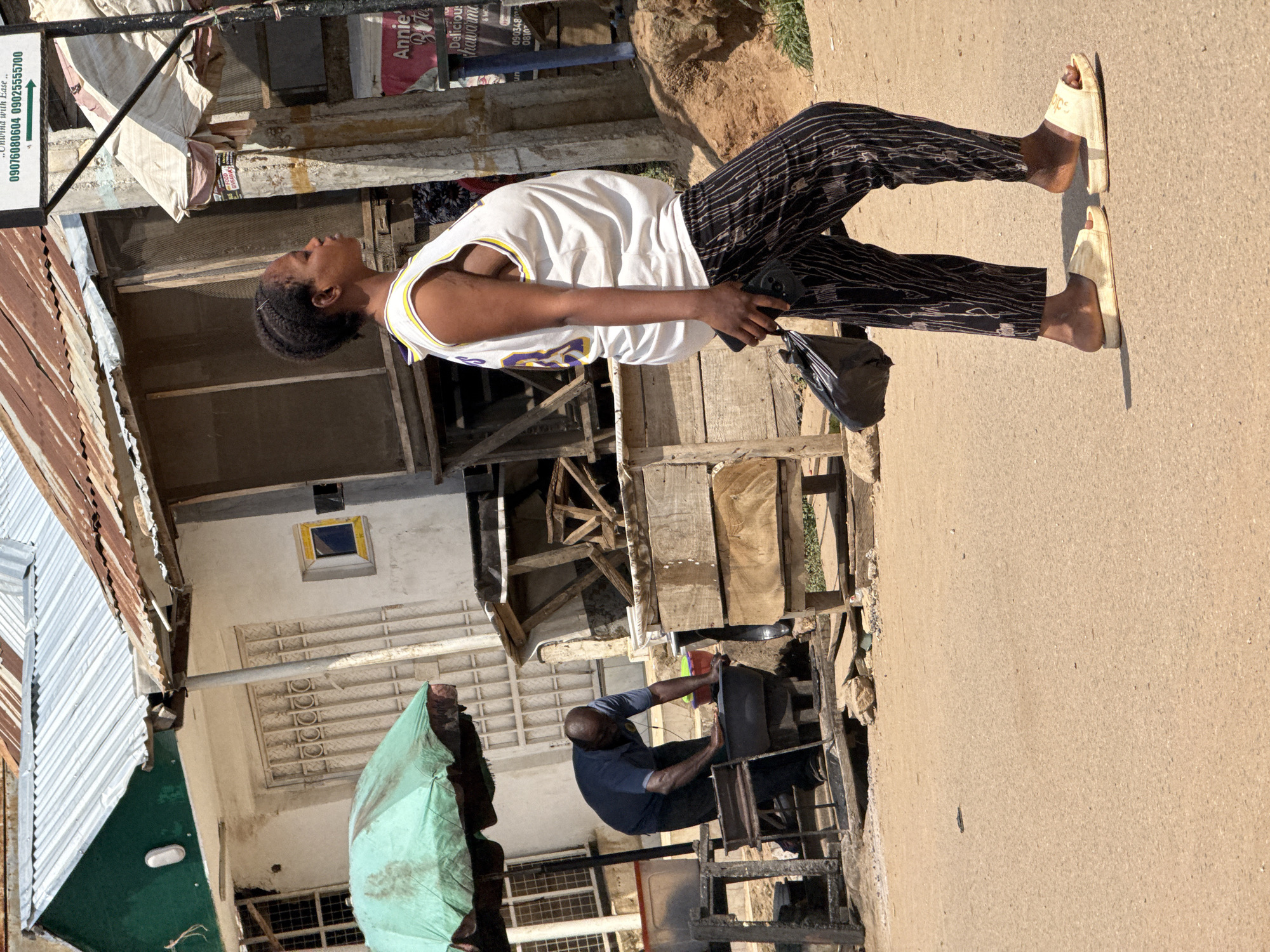 A woman walks across a dusty, unpaved street in Abuja, Nigeria, holding a black plastic bag in her right hand and a black cell phone in her left. She is wearing a white sleeveless shirt with yellow trim, black and white striped pants, and cream-colored slides. To her left, a man is bent over a large black basin, appearing to wash something. He is wearing a dark blue short-sleeved shirt and dark pants. In the background, there are several structures with corrugated metal roofs, some appearing weathered and rusty. A green umbrella-like awning is visible over the man's workspace. Signs are present on some of the buildings, with one in the upper right corner displaying phone numbers. The ground is a mix of dirt and sparse vegetation.