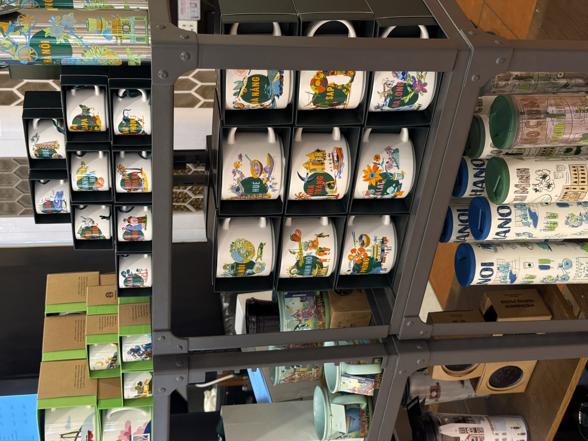The image shows a grey metal shelf unit displaying Starbucks merchandise, likely within a retail store in Đà Nẵng, Vietnam. The focus is on a selection of coffee mugs and tumblers featuring Vietnamese city names and related illustrations. The mugs, arranged in black boxes, are labelled with names like "Đà Nẵng," "Sapa," "Hạ Long," "Huế," "Nha Trang," "Đà Lạt," "Hà Nội," and "Hải Phòng." The tumblers display similar themes, with some labelled "Hồ Chí Minh" and "Hanoi." Other items, such as coffee filters, are also present on the shelves. The environment appears to be a retail space based on the organized display and presence of packaged merchandise.