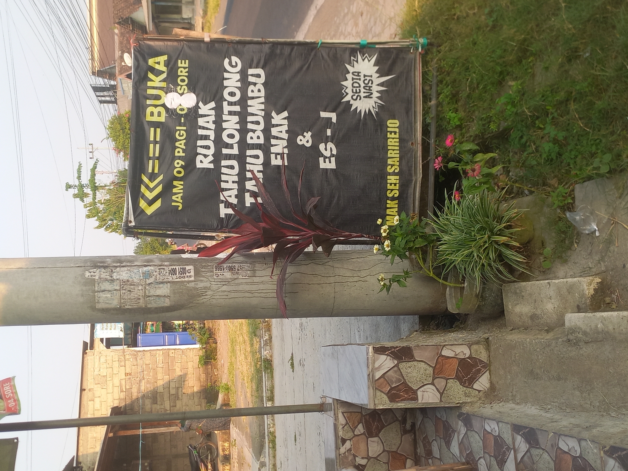 The image depicts a street scene in Pulo Gedang, Indonesia. A large black banner hangs on a pole, advertising "Rujak Tahu Lontong," "Tahu Bumbu Enak," and "Es-J." The banner also indicates operating hours: "Jam 09 Pagi - O Sore." Below the banner, smaller text reads "Mak Seh Sarirejo." To the left of the banner, a building constructed with light-colored bricks is visible, with a motorbike parked in front of it. A concrete utility pole stands prominently in the foreground, covered in various peeling posters and stickers, some of which appear to be advertisements or official notices. A concrete bench with decorative stone tiles is situated to the left of the utility pole. In front of the banner and to the right of the utility pole, various plants and flowers are growing, adding a touch of greenery to the scene. The ground appears to be a mixture of paved surfaces and grassy areas, with some small concrete steps leading down from the elevated bench area. The sky is clear and bright, suggesting it is daytime.