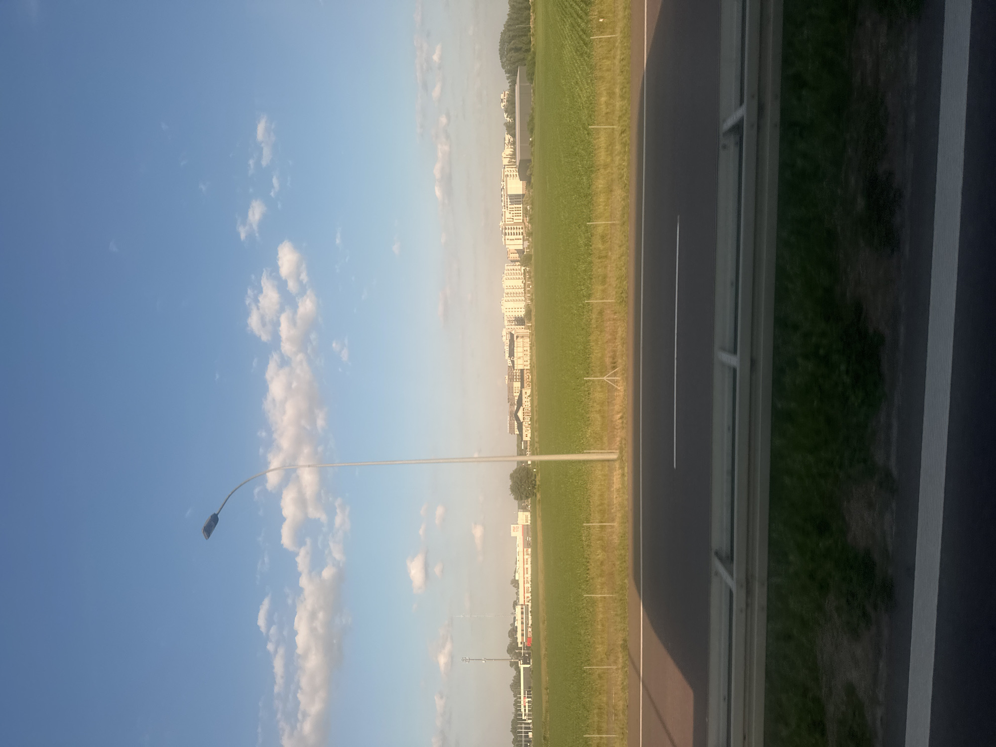 This is a vertical, eye-level, outdoor, daytime photograph of a landscape with a road in the foreground and buildings in the distance. The sky is a clear blue with scattered white clouds. A tall, grey lamppost stands prominently in the left-center of the frame.
The middle ground features a vibrant green field of crops, possibly corn, with a row of thin, vertical poles running horizontally across it. Beyond the field, a cluster of buildings forms a distant skyline. The buildings are predominantly light-colored, appearing to be residential or office structures with multiple windows. To the far left, a tall communications tower is visible, and further to the right, there are some trees.
The foreground is dominated by a dark grey asphalt road with a white line marking its edge. A silver guardrail separates the road from the grassy verge. The scene appears to be located on the outskirts of a city, possibly Garwolin, Poland, given the architectural style and agricultural landscape. There are no people or animals visible in the frame, and no actions or interactions are depicted. The overall atmosphere is serene and quiet.