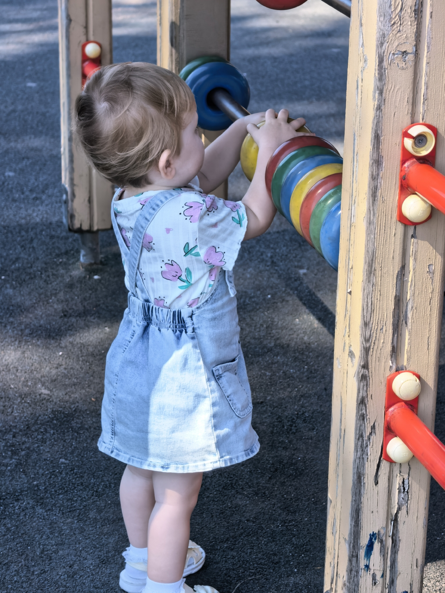 A young child, facing away from the camera, plays with a colorful abacus on a playground. The child wears a light-colored denim overall dress with a floral pattern on the top and white socks with sandals. The abacus features large, circular beads in shades of blue, yellow, green, and red. The playground structure is made of weathered wood and painted metal. The ground is covered in dark, granular material. The scene is lit by natural sunlight, casting shadows on the ground. The location is identified as Sterlitamak, Russia.