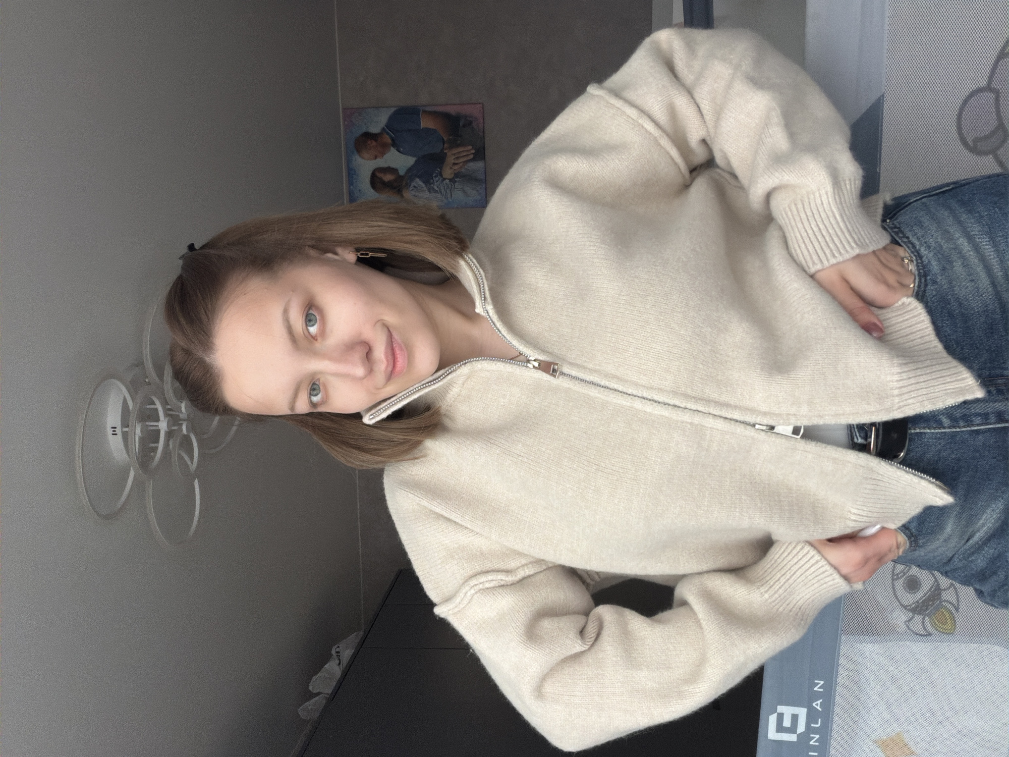 The image features a young woman posing indoors, likely in a room within a home in Reshetnikovo, Russia. She has light brown hair, styled with a small black clip, and striking blue eyes, engaging directly with the camera. Her expression is neutral to gently smiling, conveying a calm demeanor.
She is dressed in casual attire, wearing a cream or light beige-colored knitted cardigan with a full-length zipper down the front, and blue denim jeans. Gold-toned, geometric earrings are visible in her ears. Her fingernails are painted with a light polish, and a ring is visible on one of her fingers. Her hands are positioned at her waist, subtly tucked into her jeans or holding the bottom edge of her sweater.
The background reveals elements of a domestic setting. A framed picture or painting hangs on the wall, depicting two figures (a man and a woman) in an embrace. Below it, the wall is a plain grey. To the left, a modern-looking light fixture with multiple clear circular elements is mounted on the wall, and beneath it, a white object with a "III" or similar logo. A dark surface, possibly a dresser or table, is visible in the lower left. On the right side, partially visible, is a patterned mat or blanket, featuring a rocket ship design and the partially visible text "EINLAN". The overall lighting suggests a soft, ambient illumination, indicative of daytime indoors.