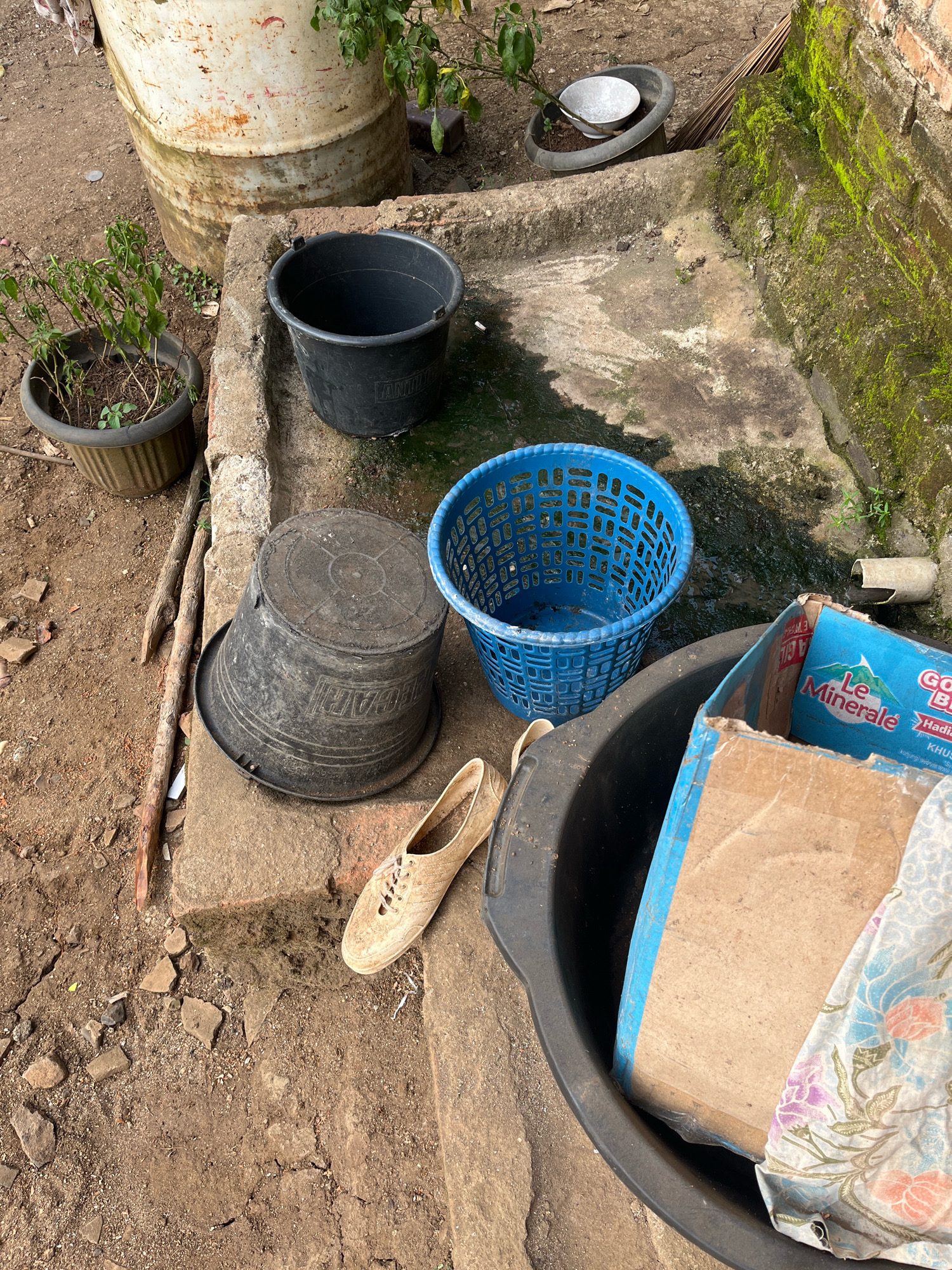 This is a photograph taken outdoors in Kebumen, Indonesia. The scene depicts a collection of discarded items and construction debris in what appears to be a neglected outdoor area. Prominently featured is a large, rusty white barrel, likely a repurposed industrial drum, situated in the background. In the middle ground, there are several plastic containers, including a black bucket, a blue plastic basket, and a large, dark gray basin. A single, worn, tan-colored shoe rests on a concrete block near the basin. A partially visible cardboard box with blue tape and a label reading "Le Minerale" is placed inside the basin, alongside a piece of fabric with a floral pattern. The ground is rough and consists of dirt, small stones, and scattered pieces of debris. To the right, a moss-covered brick wall suggests dampness and neglect. A small potted plant is visible to the left of the barrel, and some dried plant material is also present. The overall impression is one of an unkempt utility area or backyard.