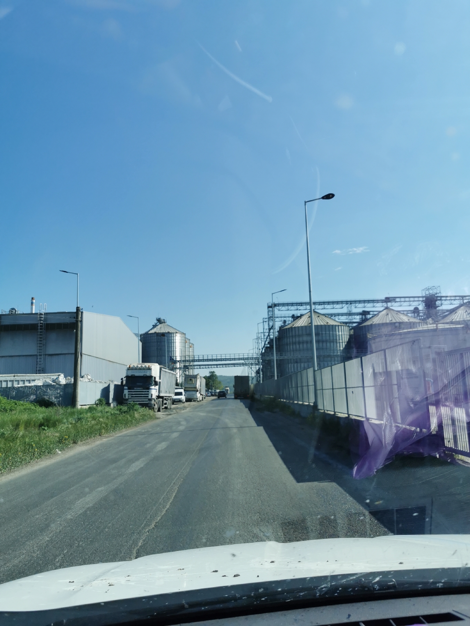 The image shows a straight road lined with industrial structures and parked trucks, possibly located in or near Razdelna, Bulgaria, given the presence of grain silos and associated facilities. The sky is clear and blue. There are several metal silos on either side of the road connected by overhead walkways. A row of semi-trailer trucks is parked on the left side of the road next to a large metal building with a ladder. Street lamps line the road. The view is taken from inside a vehicle, evident from the dashboard and windshield obstructing the lower portion of the frame. There are no people visible in the frame. The overall environment appears industrial, likely a storage or processing area for agricultural products.