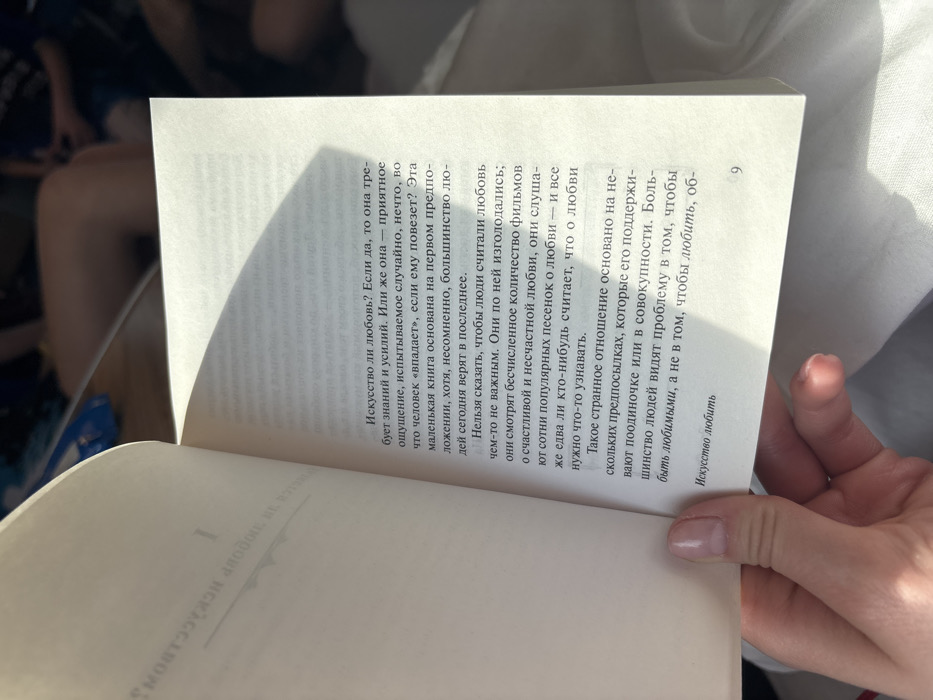 The image displays an open paperback book, identified as Erich Fromm's "The Art of Loving" in Russian (Искусство любить), held by a human hand. The scene is located in Щучье Озеро, Russia. The right page, clearly numbered "9" at the bottom, presents Cyrillic text that questions whether love is an art requiring knowledge and effort or a pleasant, accidental sensation. A prominent, sharp diagonal shadow partially covers the upper and central portions of this page. The left page shows a partial chapter title, "ИСКУССТВО ЛЮБ...", and the Roman numeral "I". The background is blurred, revealing indistinct shapes, including human legs in the upper right, and white fabric in the lower right, indicative of an indoor or enclosed setting illuminated by strong, directional natural light.