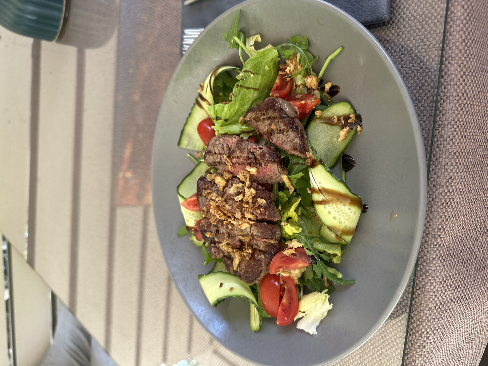 A medium shot captures a steak salad on a gray plate, positioned on a table with a textured brown tablecloth. The salad features sliced steak, lettuce, arugula, sliced tomatoes, and cucumber ribbons. Crispy fried onions are scattered over the steak. A dark drizzle, likely a balsamic glaze, adorns the salad. The background is blurred, showing a reflective surface with horizontal lines, suggesting a table or countertop. The setting appears to be an indoor dining area, possibly a restaurant or cafe. No individuals are visible in the image. The scene is brightly lit, highlighting the food. There is no discernible information in the image to indicate a specific location in Shirokaya Schel, Russia.