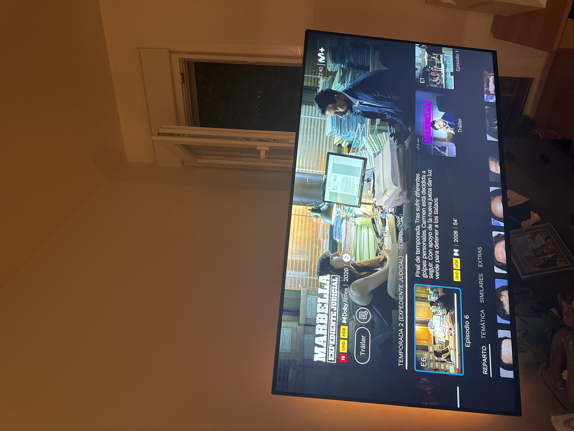 An indoor scene shows a flat-screen television displaying the streaming service interface for the Spanish television series "Marbella: Expediente Judicial." The television screen is the primary focus, occupying the majority of the frame. The series title "MARBELLA" is prominently displayed in large white letters with a red border, followed by "EXPEDIENTE JUDICIAL" in smaller white text. Below the title, various icons and information are visible, including a red "18" rating, "UHD," "HDR," "Dolby Atmos," and the year "2026." The interface shows a synopsis for "TEMPORADA 2 (EXPEDIENTE JUDICIAL)" and an episode thumbnail with "E6" and "Episodio 6" below it. The thumbnail depicts two individuals, a woman and a man, sitting at a desk with stacks of papers.
The background of the television screen shows a dimly lit office environment with bookshelves filled with binders and files. To the right of the main synopsis, there are other thumbnails for the series, including one labeled "E1" for "Episodio 1" and another for a "Tráiler." The top right corner of the television displays the date "16 JUN 22:40" and the "M+" logo.
The television is set against a backdrop of a room, with a light brown wall on the left and a white wall with a partially open window on the right. The window reveals a dark, indistinct view of the outside at night, possibly showing faint lights in the distance. The overall lighting in the room is dim, with a warm glow emanating from the television screen and a light source from the left. There are no identifiable landmarks or specific features that would indicate the city of Murcia, Spain.
