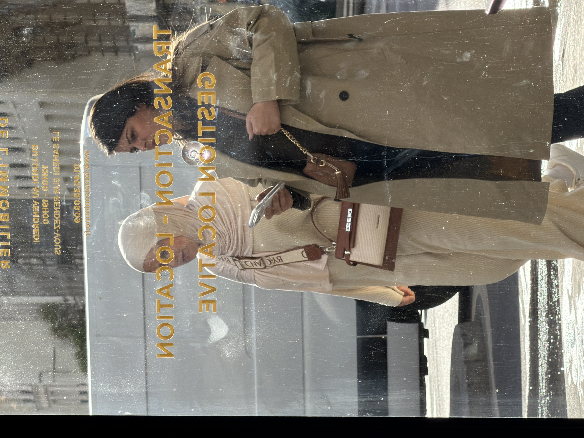 This image captures a bright, daytime street scene in Paris, France, reflected in a dusty storefront window. The clear weather is indicated by strong highlights and reflections.
Two women are visible in the window's reflection. The woman closer to the viewer wears a beige trench coat over dark clothing and carries a small brown purse with a gold chain. Further back, another woman is dressed in a light cream pleated outfit and a matching hijab. She is looking down at a smartphone held in her hand and carries a small beige and brown shoulder bag with a strap bearing the brand "BYCAND". Both women appear casually engaged, possibly walking past or pausing in front of the establishment.
The window features clear text, indicating the business is a real estate agency. The text, reflected and thus mirrored in the image but conveying the following, advertises services such as "TRANSACTION", "GESTION" (Management), and "LOCATION" (Rental). Further details include "www.lemmoniez.com", a phone number "01.40.69.09.09", availability "À VOTRE SERVICE DU LUNDI AU SAMEDI" (At your service from Monday to Saturday), and operating hours "10H00 - 19H00". Other services mentioned are "À VENDRE" (For Sale) and "À LOUER" (For Rent), all under the umbrella of "IMMOBILIER" (Real Estate).