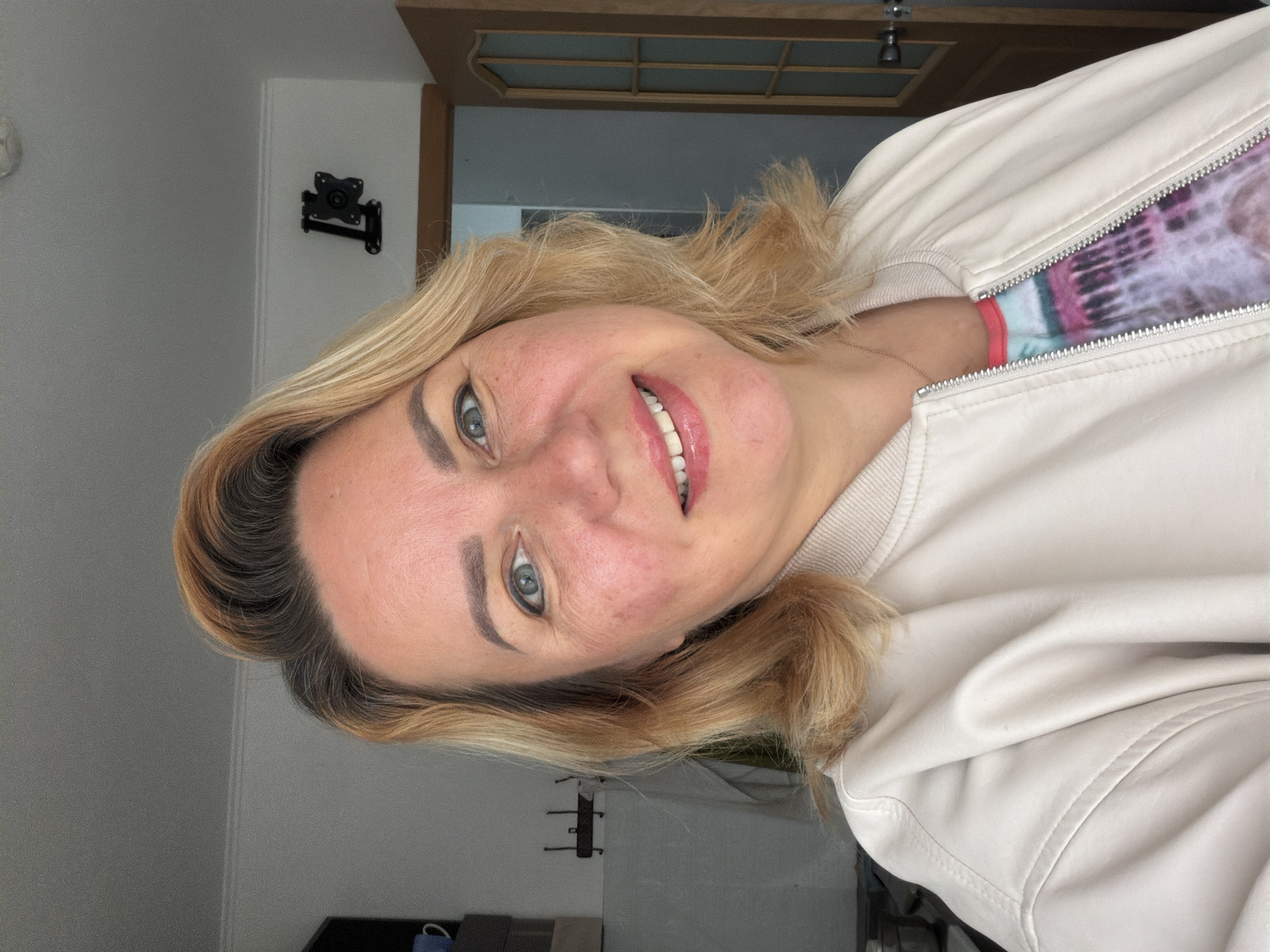 A blonde-haired woman is smiling at the camera. She is wearing a white jacket, and a tie-dye shirt can be seen underneath. The background includes a door, a wall with a bracket mounted on it, and a glimpse of flooring. The photograph appears to have been taken indoors and rotated 90 degrees. There is no information present in this photo to indicate this picture was taken in Leningradskaya, Russia.