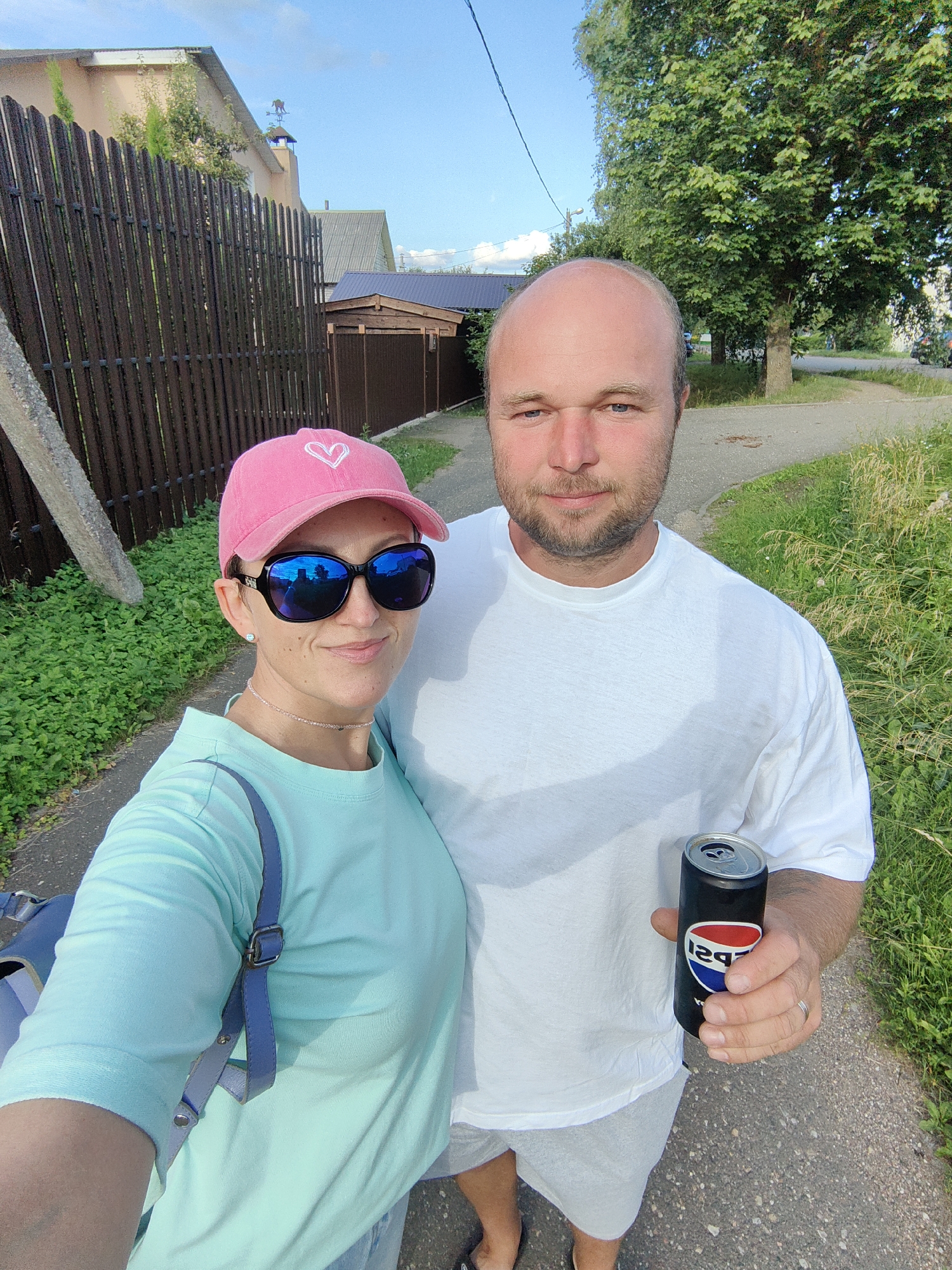 A selfie taken outdoors shows a man and a woman standing next to each other. The woman, on the left, is wearing a pink baseball cap with a white heart on it, sunglasses, and a turquoise sweatshirt. She has a light-colored necklace and a blue strap from a bag visible on her shoulder. The man, on the right, is wearing a white t-shirt and light-colored shorts. He has a beard and is holding a black Pepsi can. They are standing on a gravel path, with green grass and foliage on either side. In the background, there is a brown wooden fence, houses, and trees under a blue sky with some clouds. The location is identified as Fanipal, Belarus.