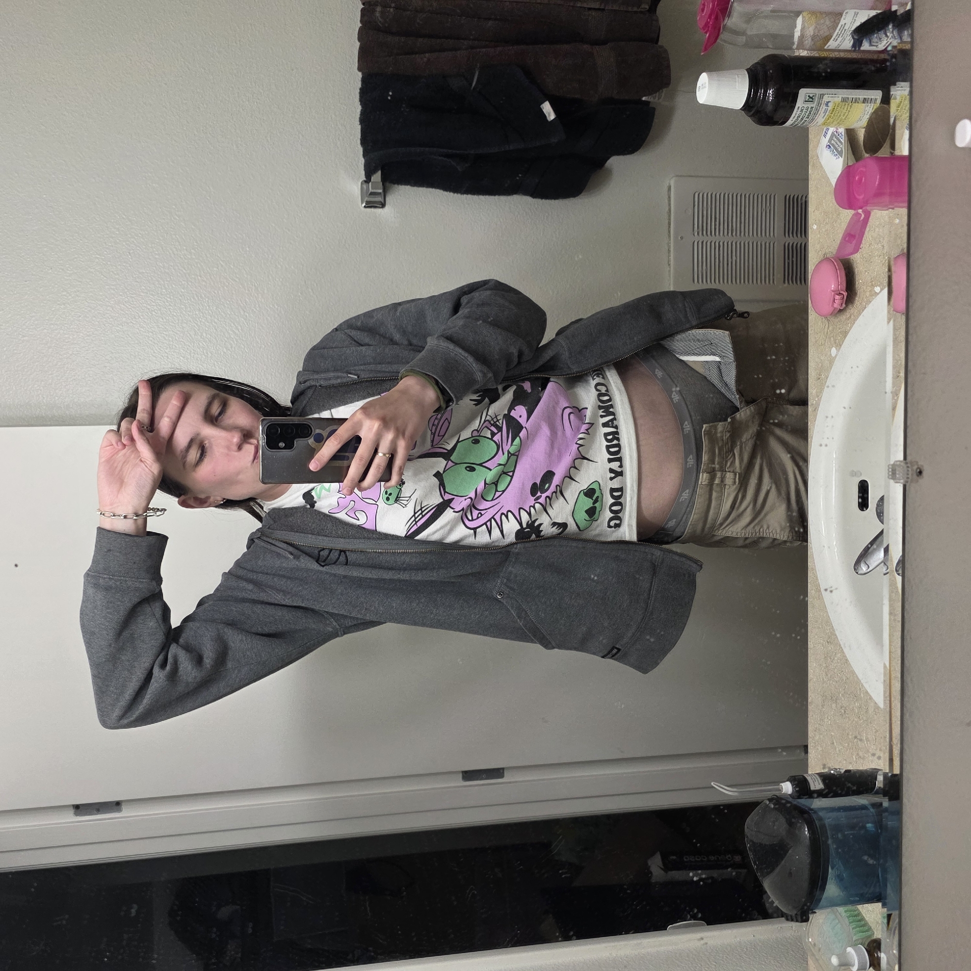 The image is a vertical, full-body mirror selfie taken in a bathroom. The subject, who appears to be a young person, is wearing a grey zip-up hoodie over a white t-shirt with a cartoon graphic and the text "COMBOY DOG" printed on it. They are also wearing khaki pants, with grey boxer briefs visible above the waistband. The subject is holding a smartphone in their right hand, using it to take the picture. Their left hand is raised to their forehead, and they are making a peace sign with their fingers. Their eyes are closed, and their mouth is puckered, suggesting a playful or coy expression.
The bathroom setting includes a white sink with a faucet, a tiled countertop with various toiletries and a bottle of dark liquid, and a wall with a towel rack holding folded towels. A vent is visible on the wall. The lighting suggests it is daytime, though the interior setting makes it difficult to determine the weather. The reflection in the mirror shows a dark area below the subject, possibly a window or another part of the room. The overall mood is casual and personal. The text on the t-shirt is clearly legible as "COMBOY DOG".