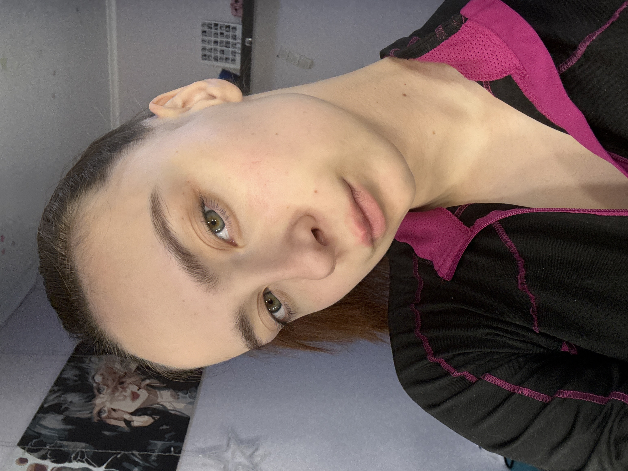 A close-up shot shows a young woman with fair skin and green eyes. She has light brown hair pulled back. She is wearing a black athletic jacket with pink trim. The background appears to be a room with light-colored walls and various items, including a poster or photo collage. There's also an artwork visible on a black and white surface. The orientation suggests the photo was taken at an angle. There is no information to suggest the image was taken in the city of Прикамский, Russia.