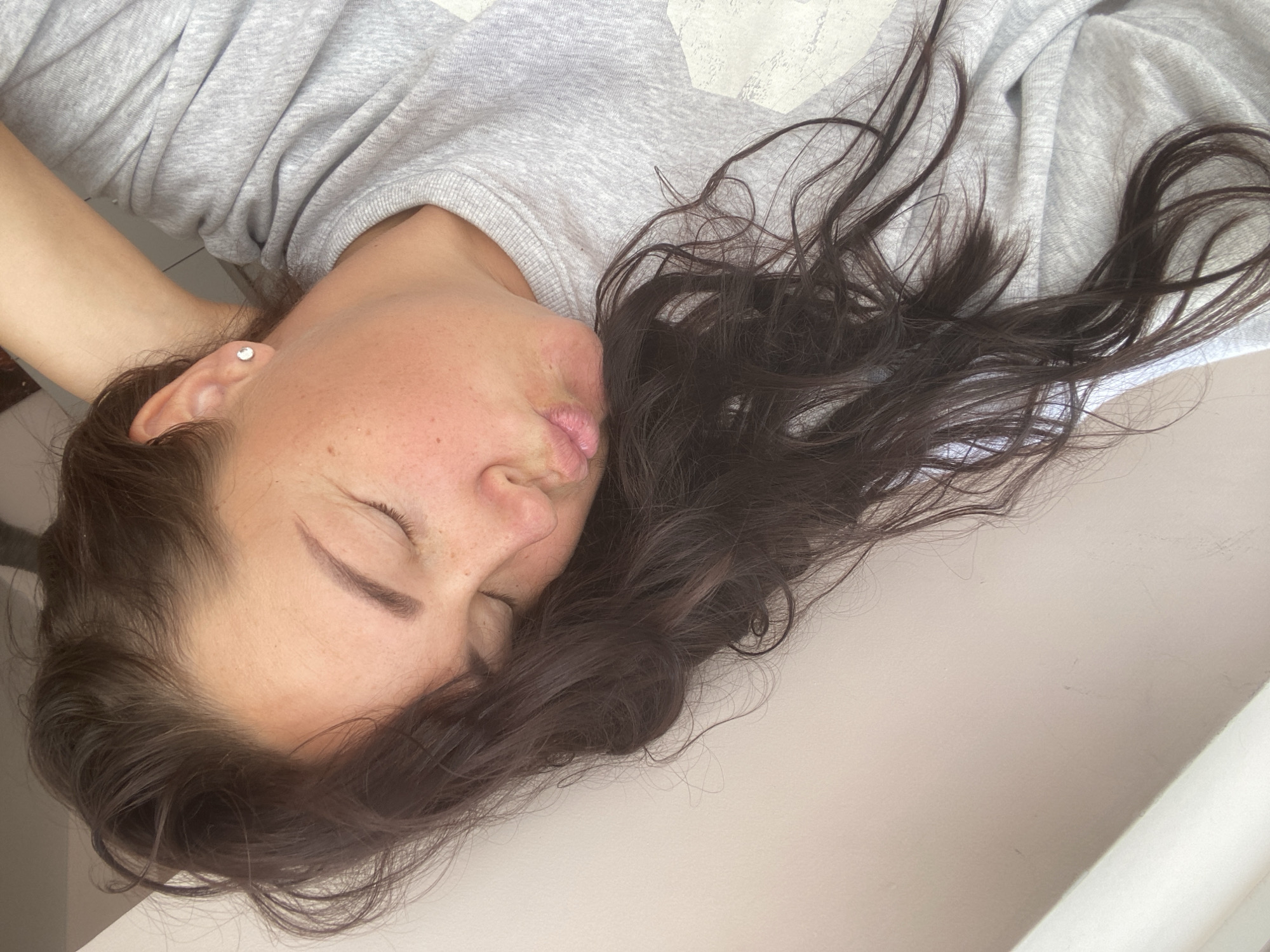 A young woman with dark brown hair is lying down and looking at the camera with her eyes closed and her lips puckered in a kiss. She is wearing a gray sweatshirt and a small diamond stud earring in her left ear. Her right arm is behind her head. The background appears to be a light-colored wall and a white surface. The image does not provide any information about the location in the city of Sochi, Russia.