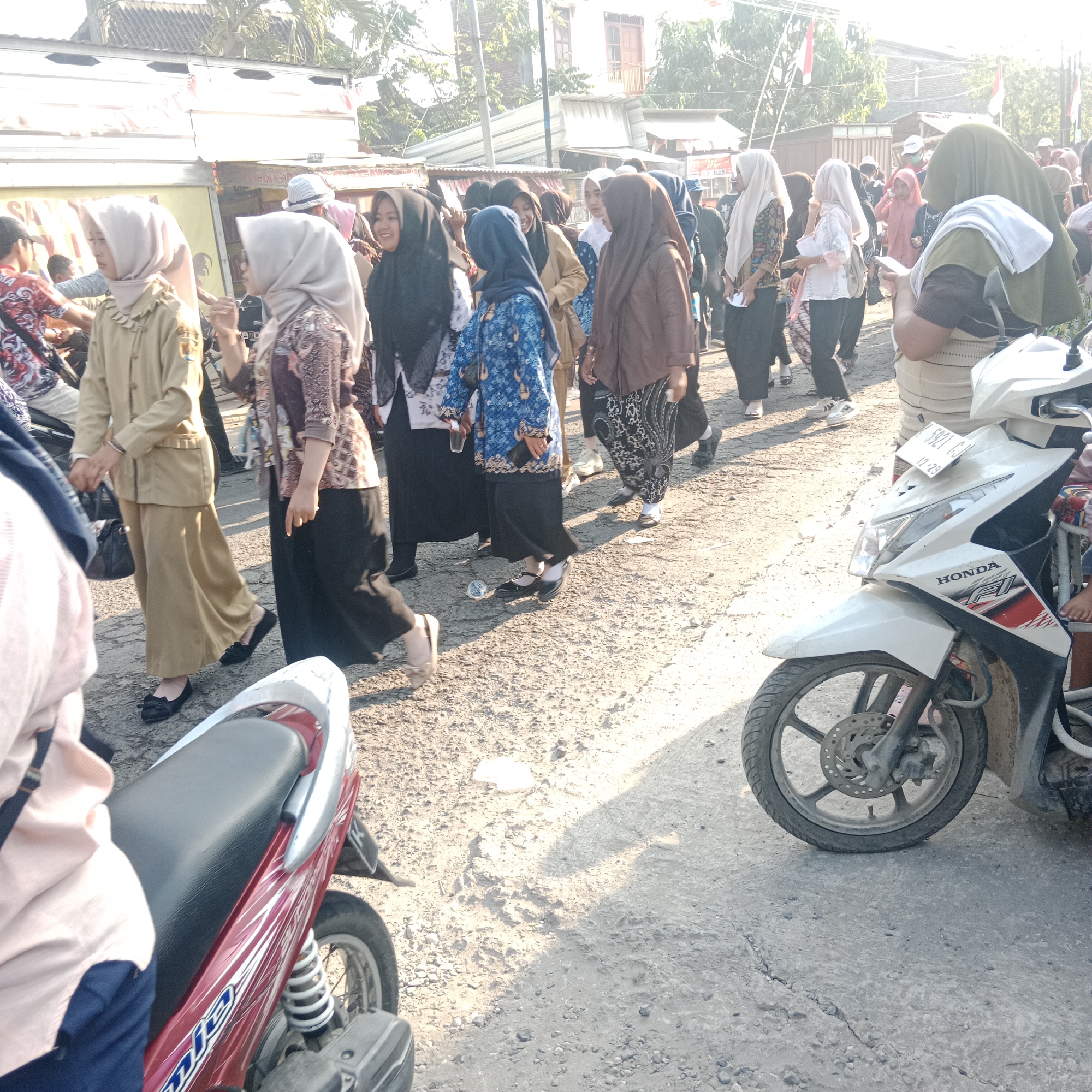 A crowd of people, predominantly women in hijabs, are walking along a dusty, cracked asphalt road in what appears to be a rural or suburban area of Tegalrejo, Indonesia. The sun is bright, casting long shadows across the ground. Several motorcycles are parked on the side of the road, with one white scooter in the foreground prominently displaying a "HONDA" logo and a license plate that reads "B 5021 CJ". The people are dressed in a variety of clothing, with some wearing uniforms that appear to be from a school or government institution, indicating a possible organized event or gathering. The buildings in the background suggest a commercial or residential street, with some shops or stalls lining the road. The overall atmosphere seems to be one of movement and activity, possibly a procession or a market day.