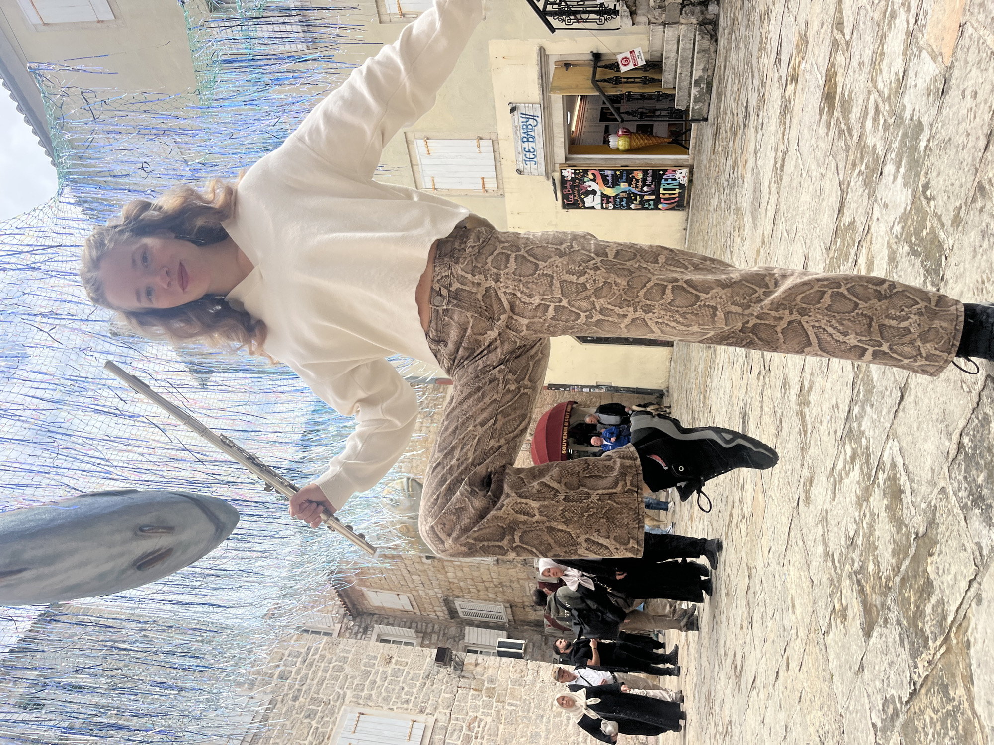 A full-body shot shows a young woman in mid-air wearing a white sweater and snake-print pants, holding a flute, with Budva, Montenegro, in the background. A decorative fish hangs from iridescent tinsel over a stone street crowded with tourists. In the background, a souvenir shop reads "Ice Baby". The orientation of the image is skewed, making the scene appear tilted.