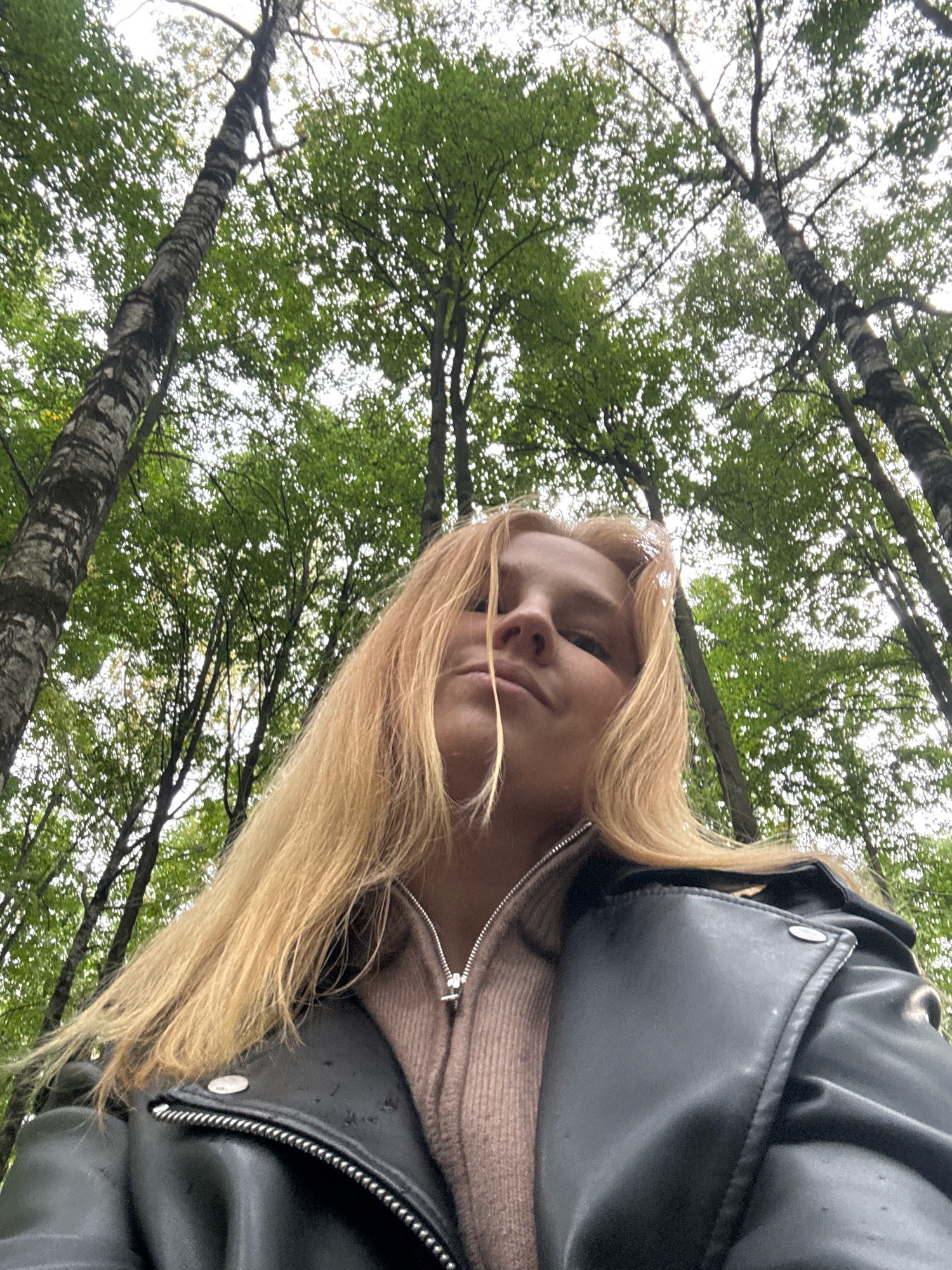 An outdoor, low-angle, waist-up shot features a young Caucasian woman with blonde hair, wearing a pink sweater and a black leather jacket. She is looking upwards and slightly to her right. The background consists of a dense forest with tall trees and green foliage against a bright, overcast sky. There is no indication of the city of Yaroslavl, Russia, in the image.