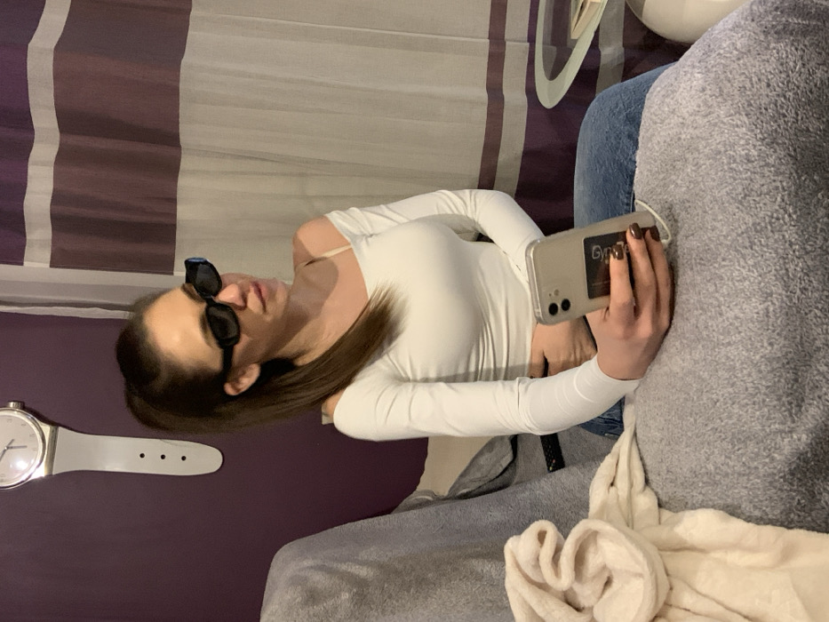 A single adult female is seated indoors within what appears to be a commercial establishment. She wears a white off-the-shoulder long-sleeved top, blue jeans, and rectangular black sunglasses, with her medium-brown hair styled in a high ponytail. She holds a light-colored smartphone, featuring a dual camera system and a dark sticker partially legible as "Grrr...", in her right hand. Her fingernails are painted a dark shade. She is looking towards the right side of the frame, away from the viewer.
The subject is seated on grey upholstered furniture, with a cream-colored folded towel or blanket placed to her left. A black remote control is partially visible on the upholstery behind the towel. The background features a plain purple wall on the left, adorned with a large, white, watch-shaped wall clock that includes a strap-like extension with three simulated holes. To the right, translucent white curtains with horizontal purple stripes hang, partially obscuring a clear glass table and a white, rounded object. The lighting appears to be ambient indoor illumination. The precise location within Sofia, Bulgaria, is not identifiable from the visual information provided.