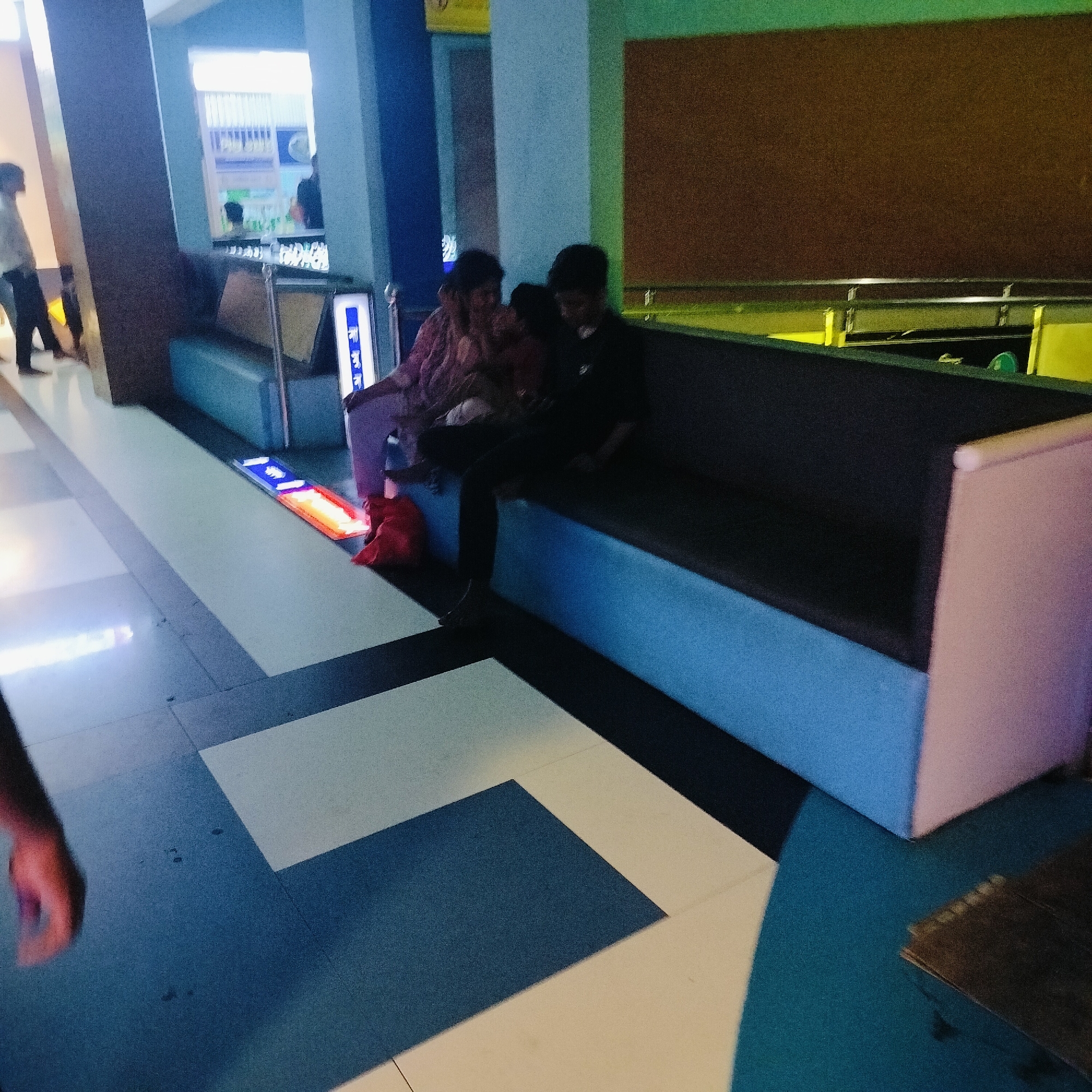 The scene shows an interior setting in Dhaka, Bangladesh, featuring people seated on a long bench. The flooring is tiled in a pattern of blue and white squares. The bench is two-toned, with a dark upper cushion and a light blue base. A woman in a pink shirt is seated next to a child. A young man in dark clothes sits to their right. A red bag is on the floor near the woman. A pair of people are walking in the background. The lighting appears to be artificial.