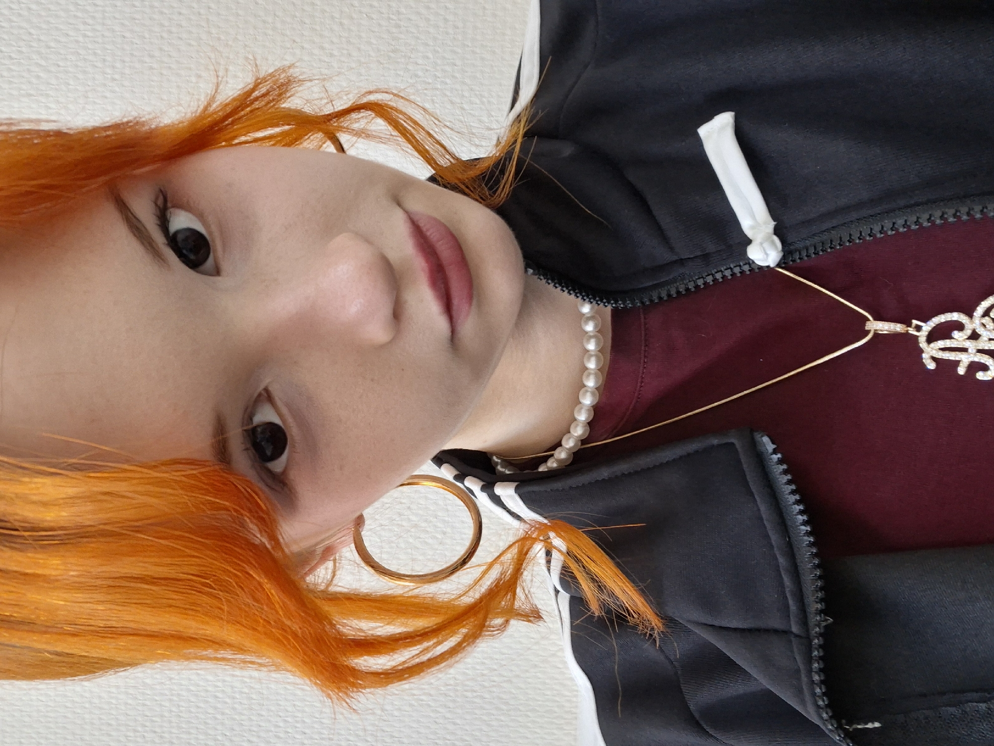 A fair-skinned person with orange hair is depicted from a close, tilted perspective. The person wears a black jacket with white stripes, a maroon shirt, a gold chain with a rhinestone pendant, a pearl necklace, and a gold hoop earring. The background appears to be a white surface. The location is not discernible from the image.