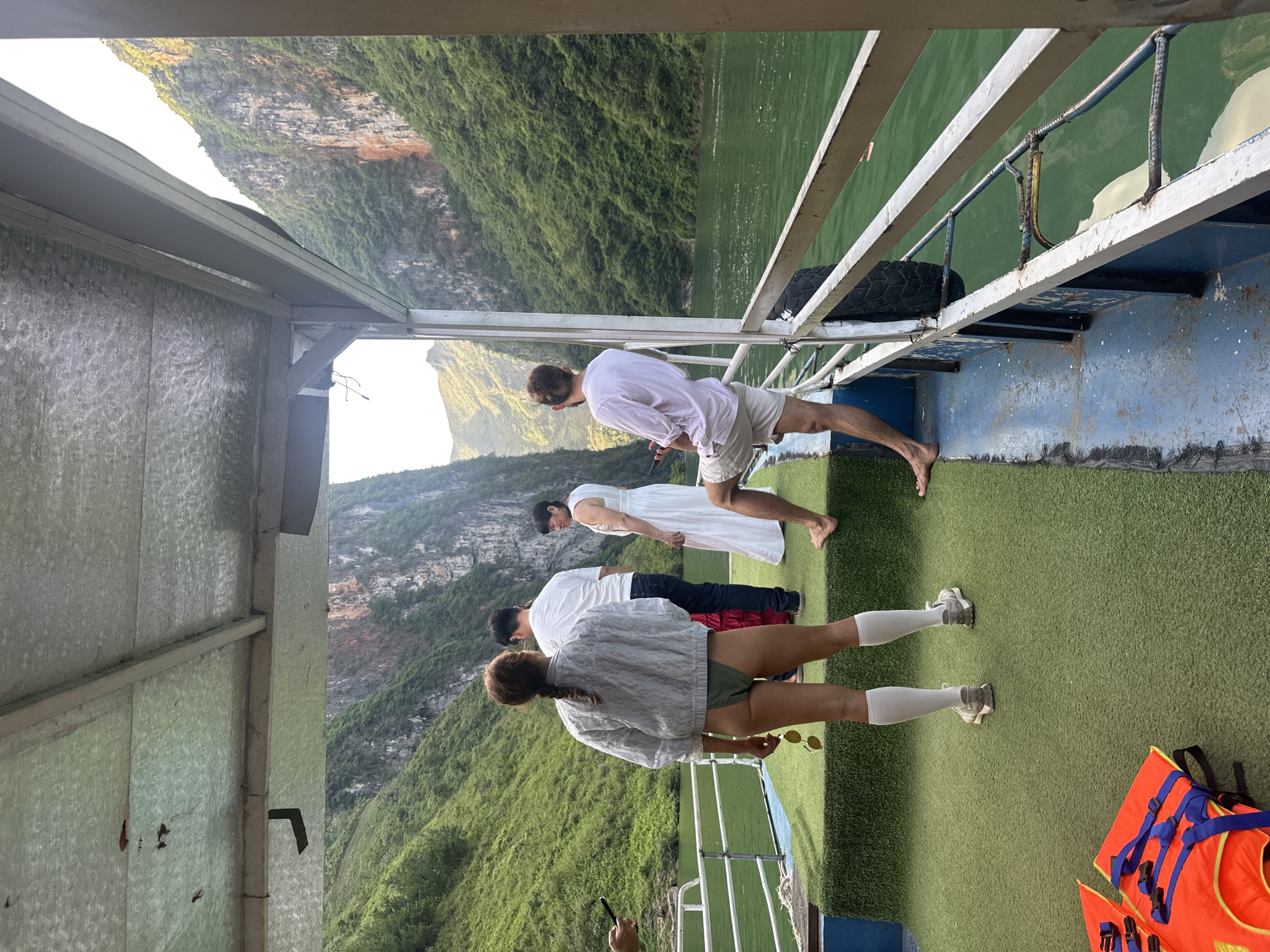 On what appears to be a boat, four individuals are gazing over the edge, likely observing the view of the surrounding environment. In the background is a view of a mountain range covered in green vegetation, typical of Southeast Asian landscapes. The image is taken from a slightly elevated position, looking down on the group of people. The image depicts a boat scene on a river, with the mountains visible in the background, likely in Đồng Văn Commune, Vietnam.