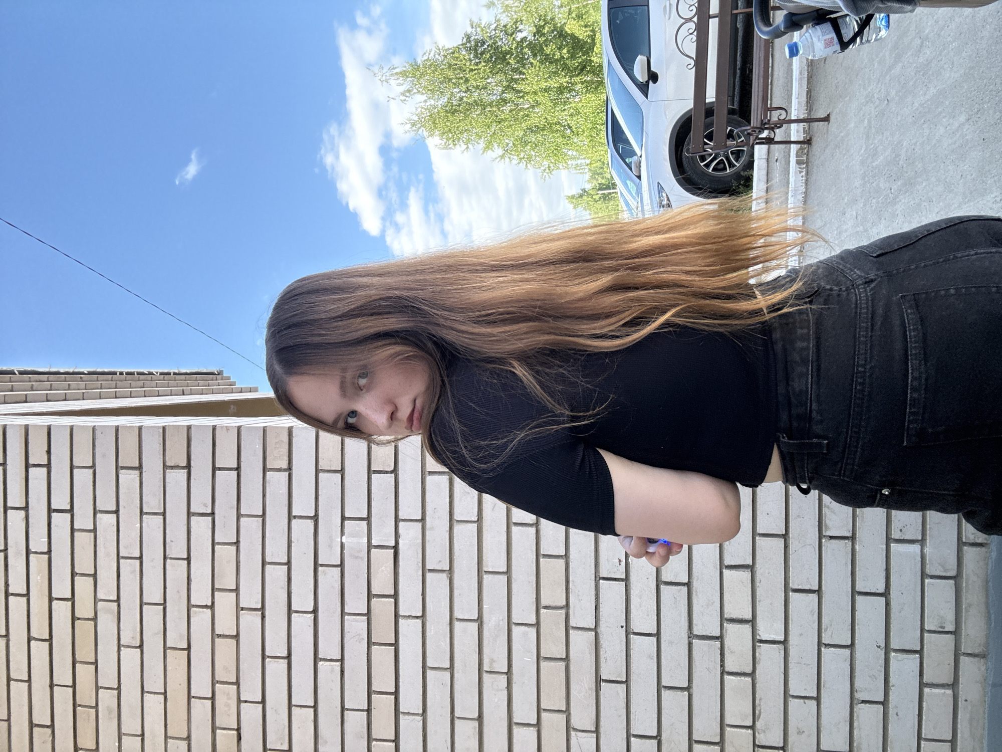 The image captures a young female outdoors in Chaikovsky, Russia, positioned with her back primarily facing a light-colored brick wall on the left. She turns her head and upper torso to look directly at the camera over her left shoulder. She has long, light brown hair and is dressed in a black short-sleeved ribbed top and black denim jeans. Her left arm is bent, holding a translucent object that emits a distinct blue glow. The background on the right reveals a clear blue sky with scattered white clouds, bisected by a thin dark cable stretching diagonally across the upper right. Green deciduous trees are visible in the mid-ground, beyond which two white SUVs are parked. A dark metal bench with ornate scrollwork is situated on a concrete or paved surface in the lower right, adjacent to a plastic water bottle with a blue cap secured in a black holder. The scene is illuminated by natural daylight.