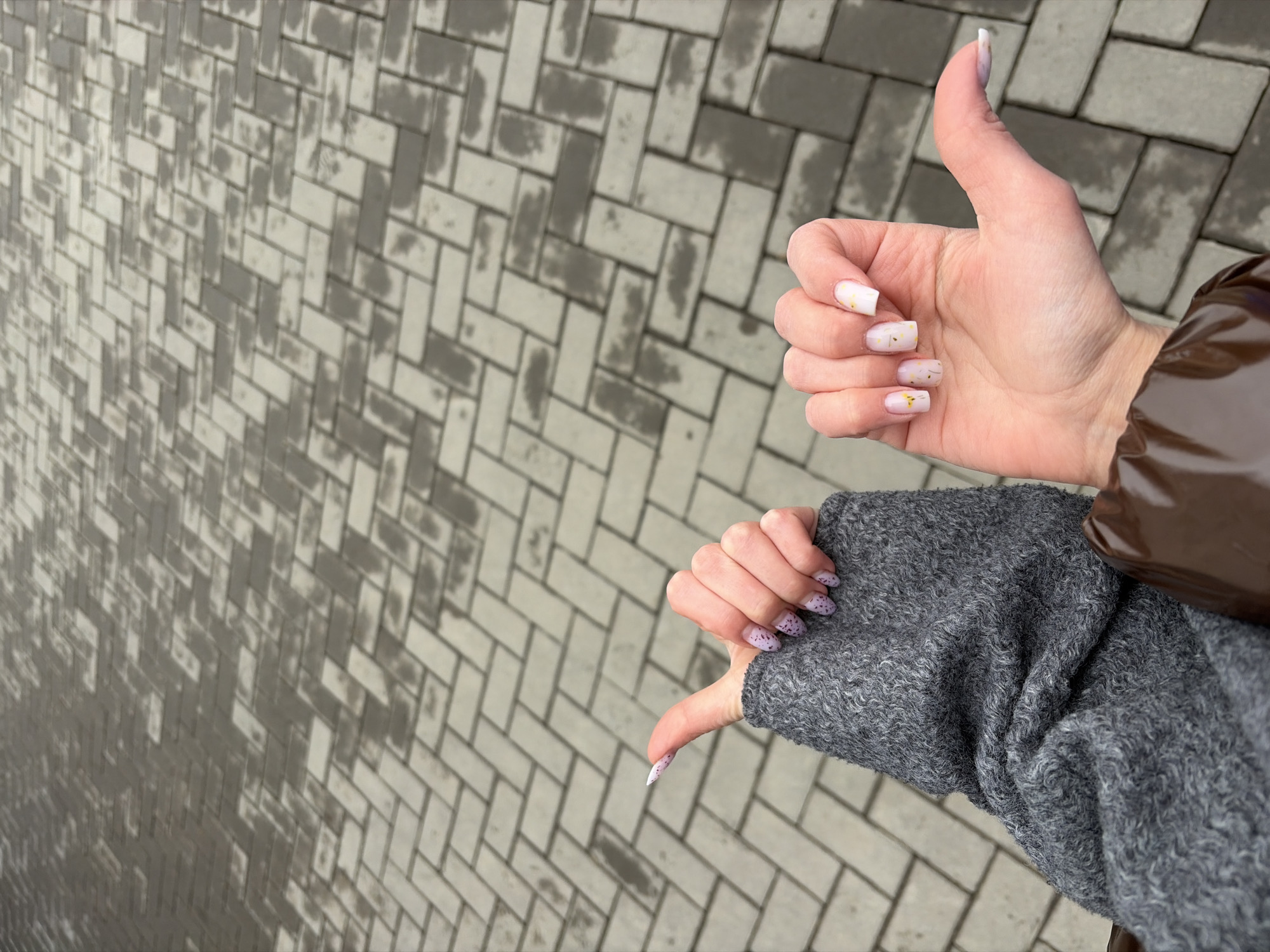 A person gives mixed feedback via hand gestures. One hand gives a "thumbs up," and the other gives a "thumbs down." The "thumbs up" hand has short nails painted with white nail polish and tiny gold stars, with a puffy brown sleeve behind it. The "thumbs down" hand has short nails painted with a white base and purple dots. This arm is covered by a puffy gray sleeve. The background is a gray brick surface. The photo was taken outside on a cloudy day. The location is likely Izhevsk, Russia.