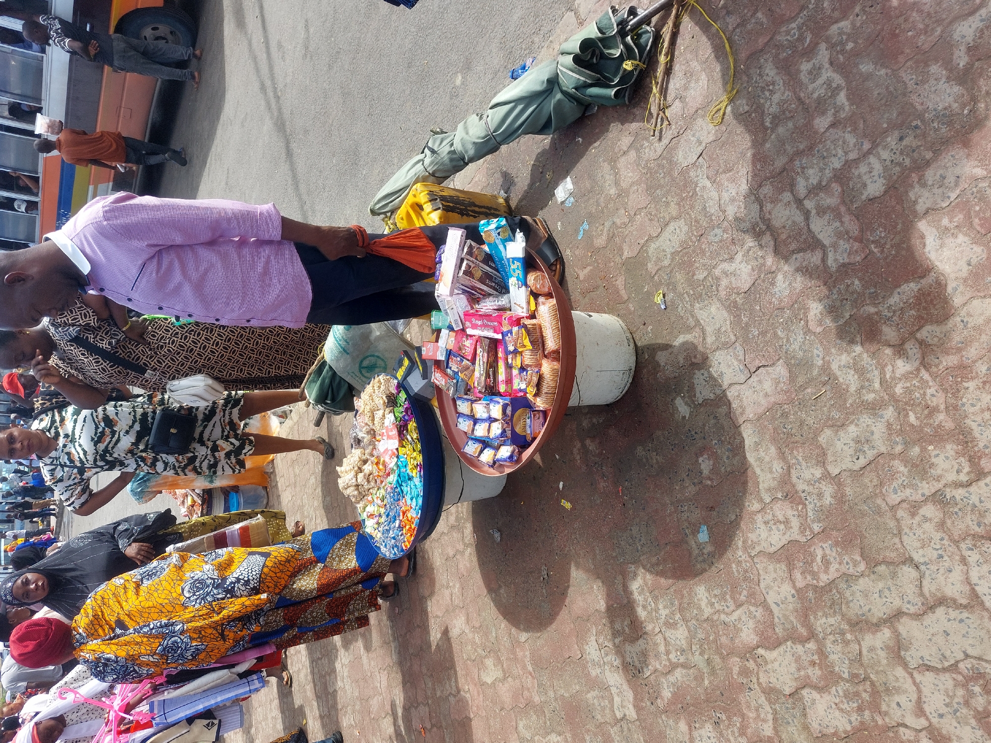 The image depicts an outdoor street scene in Dar es-Salaam, Tanzania, characteristic of a busy market or transit area such as Kariakoo. The time is daylight, indicated by pronounced shadows.
The foreground is dominated by a reddish-brown interlocking brick pavement. Two circular vendor trays, each resting on a white bucket, are positioned on this pavement. The left tray, blue, contains an assortment of colorful wrapped candies and small snacks. The right tray, brownish, holds various packaged biscuits, chocolates, and cigarettes. To the right of these trays, a green rolled-up umbrella secured with yellow string lies on the pavement alongside several wooden sticks. A yellow container is also present.
Standing on the pavement from left to right:
1. A Black female, seen from behind, wears a vibrant yellow, blue, and white patterned dress with a red head covering. She holds multiple pink plastic hangers displaying clothing items.
2. A Black female in a black hijab faces forward.
3. A Black female in a white dress with black tiger-like stripes, carrying both a black cross-body bag and a white shoulder bag, stands with hands on her hips, looking to the left.
4. A bald Black male, wearing a light purple collared shirt, dark trousers, and dark sandals, stands looking downward. He holds a small orange bag in his right hand. A green canvas bag is at his feet.
In the background, an asphalt road runs parallel to the pavement. A blue and orange public minibus (daladala) is visible on the road, with passengers inside. Several other pedestrians are present, including a man in an orange shirt walking away from the camera near the bus, carrying a clear plastic bag. The pavement surface shows wear and minor debris.