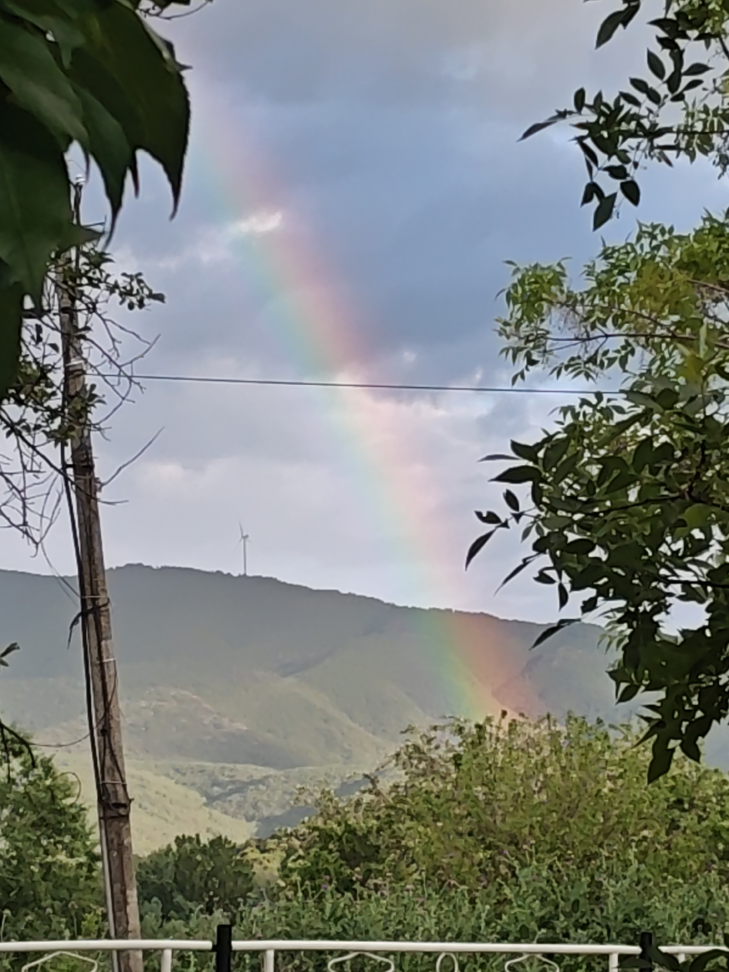 The image captures a view of a rainbow extending from the sky to the ground, framed by vegetation and a mountain range. A wind turbine is visible on one of the mountains. An electrical pole is to the left side of the image. A white fence is in the foreground. The location is Unknown, Unknown.