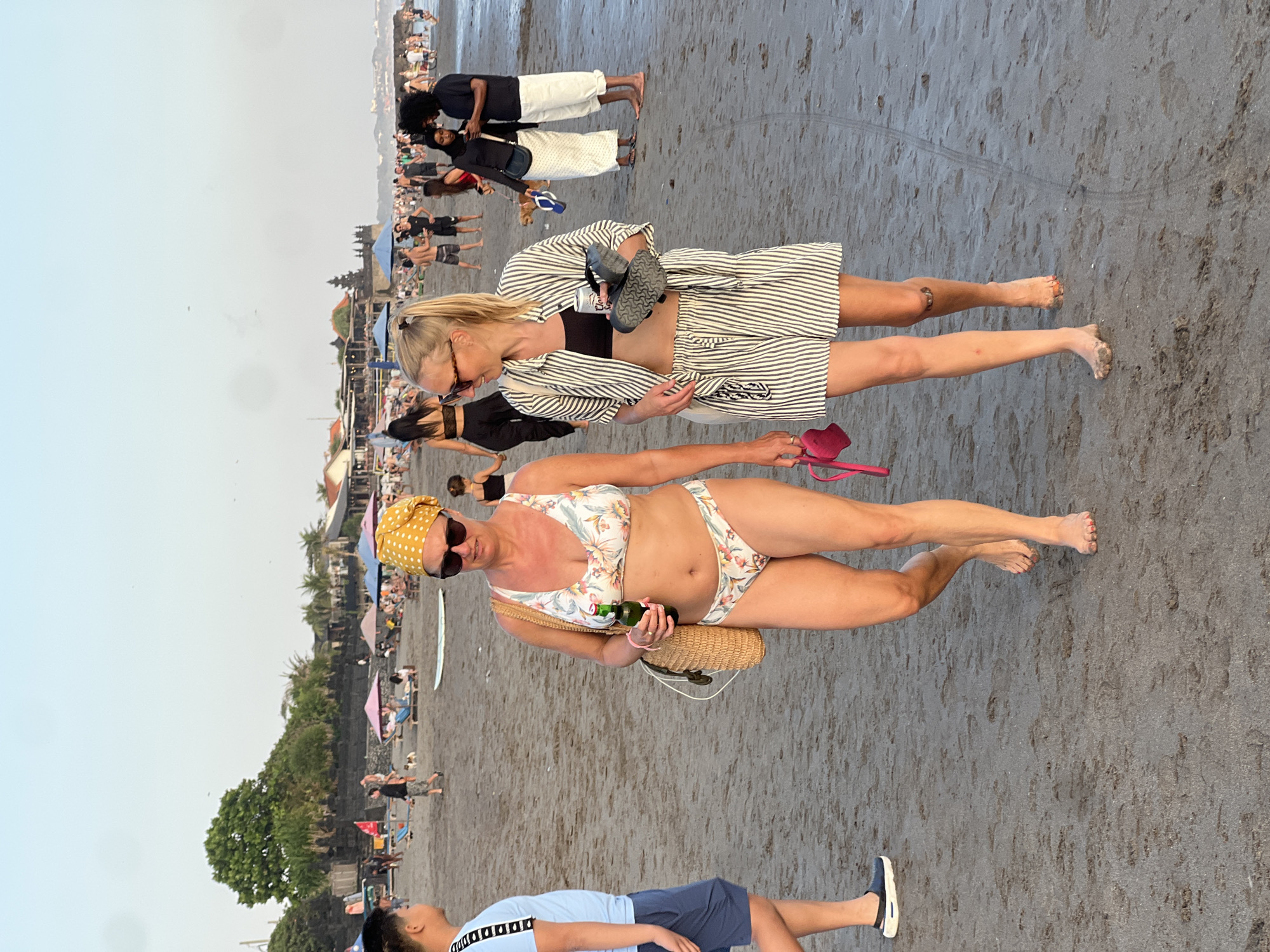 Two women are walking on a sandy beach in North Kuta, Indonesia. The woman on the left is wearing a floral bikini and a yellow headscarf, and is holding a bottle of beer and a straw bag. The woman on the right is wearing a black crop top, a striped shirt and shorts set, and is carrying flip-flops. They are walking towards the viewer. In the background, there are other people on the beach, umbrellas, and some buildings. The sky is clear and light.