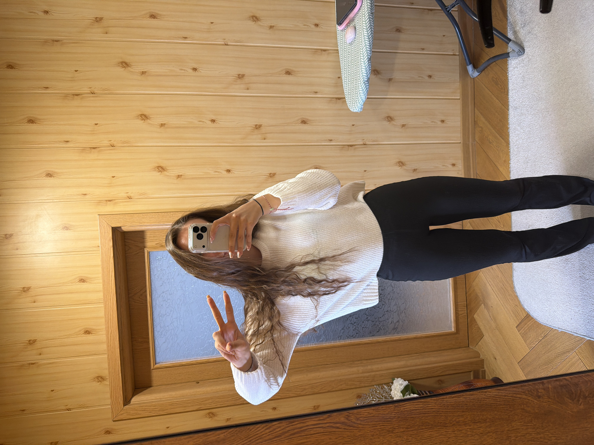 A person is reflected in a large mirror, taking a selfie. The individual wears a white sweater and black leggings, holding a cell phone up to their face. They are making a peace sign with their hand. The background includes a wooden-paneled wall, a chair, a rug, a wooden floor, and a floral decoration. There is a window in the reflection with translucent glass. No information indicates the city of Buhovo, Bulgaria.