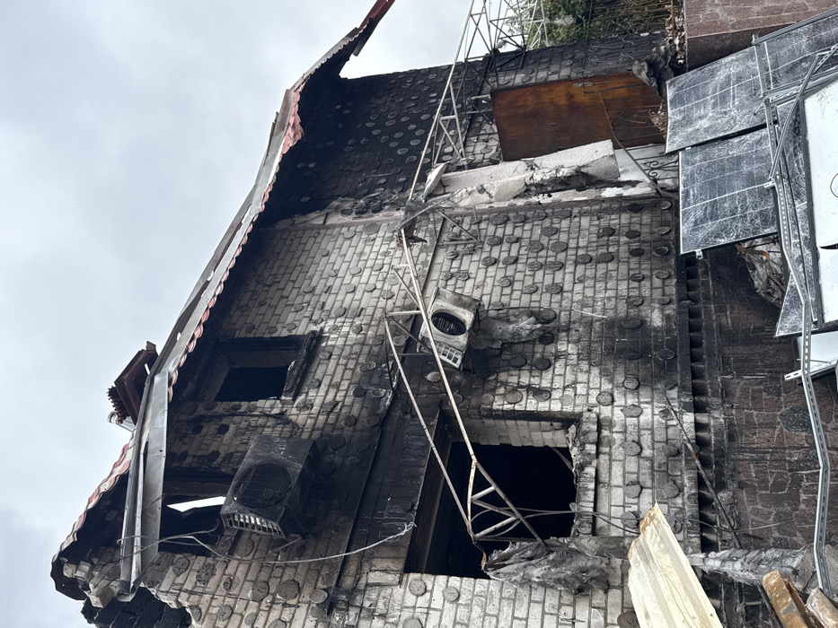 A severely damaged building is shown, possibly due to fire or explosion. The brick facade is heavily marked with soot and charring. Windows are broken or missing. An air conditioning unit hangs precariously from the wall. A ladder leans against the structure, possibly used for access or escape. The roof is partially collapsed with exposed metal and broken tiles. Debris is scattered around the building. The sky is overcast.
