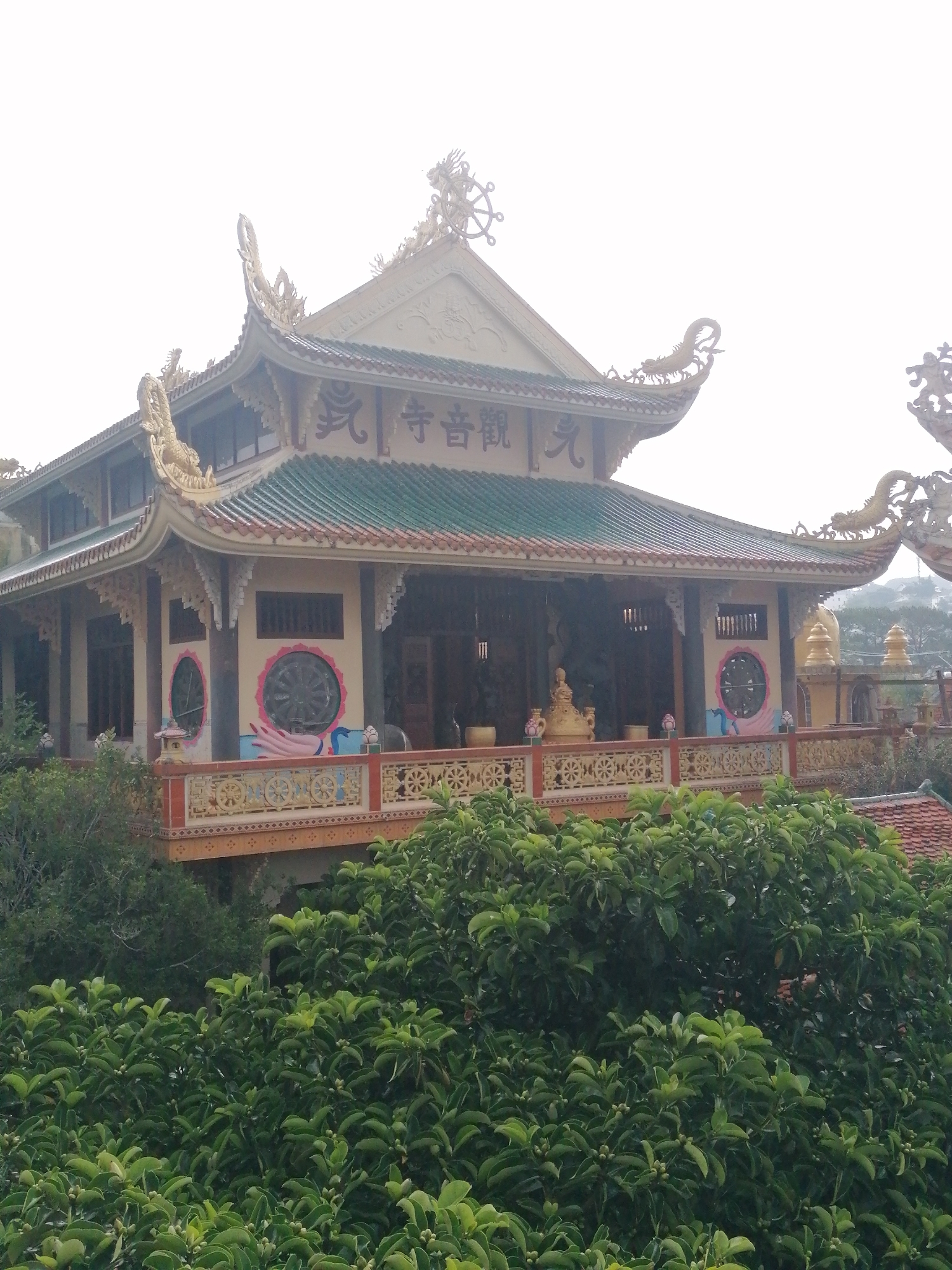 The image displays a multi-tiered Buddhist temple, identified as Quan Am Pagoda (觀音寺) from the visible text on its facade, set against an overcast sky in Da Lat, Vietnam. The temple exhibits traditional East Asian architectural elements, featuring a prominent dark green tiled roof with gracefully curved eaves, elaborately decorated with gold-colored ornamental dragons and dharma wheels.
The building's main section is a light beige color, featuring an elevated balcony with a detailed railing adorned with circular motifs. On the lower level of the facade, two large circular window-like designs are visible, one notably depicting a painted hand. Golden Buddha statues are discernible within the open interior of the temple.
The foreground is filled with a dense canopy of lush green trees and bushes, partially obscuring the temple's base and suggesting its elevated position on a hillside. In the distance to the right, faint outlines of hills or mountains can be seen. The sky is bright and hazy, indicating daytime with diffused light. No people are visible, and no specific activity or event is taking place, presenting a serene architectural view.