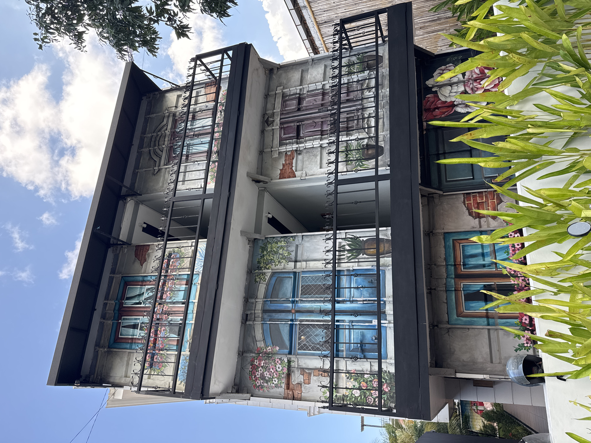 This is a full-frame, eye-level exterior shot of a three-story building with a mural painted on its facade, located in North Kuta, Indonesia. The mural depicts various windows with ornate iron grills and flower boxes, creating an illusion of depth and architectural detail. The building has multiple balconies with black metal railings. The sky is visible in the upper portion of the image, with scattered clouds and a few trees on the right side. In the foreground, lush green foliage partially obscures the view of the lower part of the building, where a motorcycle helmet rests on a white ledge. The mural artwork is detailed and colorful, featuring elements like bricks, potted plants, and draped curtains. The overall impression is that of a building adorned with trompe-l'oeil artistry.
