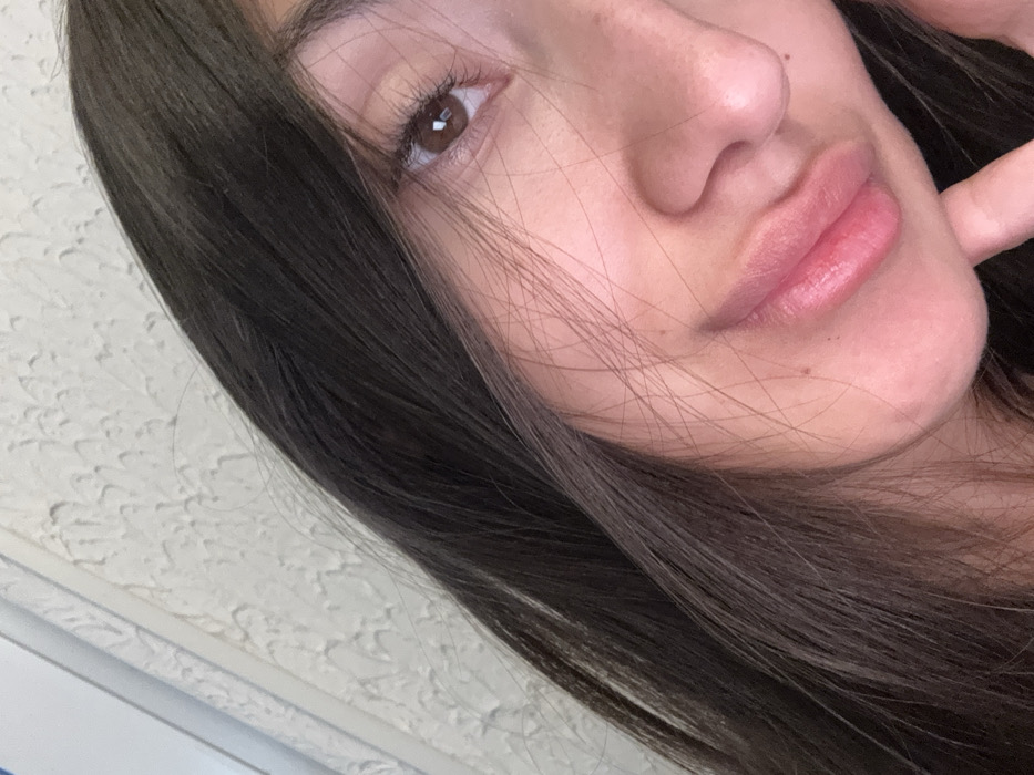 The scene is located in Zankovići, Montenegro. The image captures an angled, close-up view of a single individual, predominantly showing the right side of their face. The subject possesses long, straight, dark brown hair that partially covers the upper left portion of the frame and some facial features. The visible right eye is brown, with distinct lashes, and is directed towards the camera. The nose and mouth are visible, with the lips exhibiting a pink tone and appearing slightly parted. A finger from the subject's right hand is positioned against the skin below the lower lip. The background consists of a light-colored, textured wall surface, potentially stucco, with a narrow, slightly darker vertical element visible on the far left. The lighting is uniform, illuminating the subject's face.