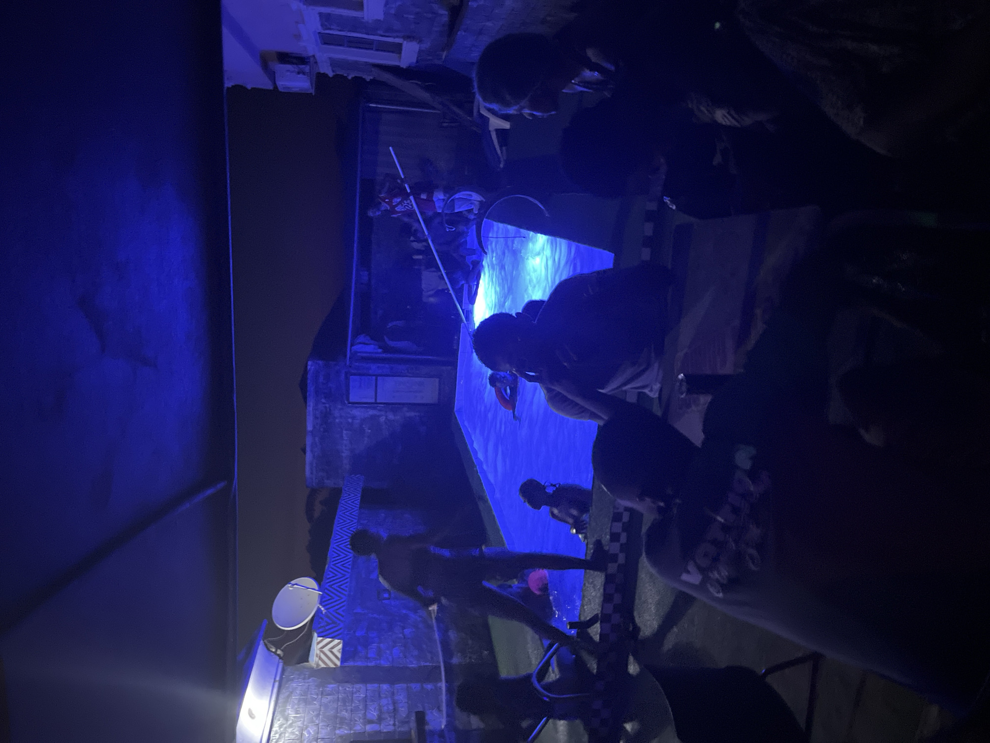 This nighttime image captures an outdoor social gathering centered around a bright blue-illuminated swimming pool, at an unknown location within the city of Unknown, Unknown. The scene is bathed predominantly in a vibrant blue light, giving a cool, ethereal cast to the entire environment and rendering many details in silhouette or shades of blue.
In the mid-ground, a rectangular swimming pool glows intensely blue from internal lighting. Several individuals are in or around the pool. On the left side, one person with dark skin is visible near the pool steps. Another individual, with lighter skin tone, stands on the pool edge, facing right, holding a white hose or pipe. Further right in the pool, a partially submerged person with lighter skin tone faces the viewer. In the deeper end, another person appears to be wearing a red flotation device. On the far right bank of the pool, a person in a red basketball jersey with the number "23" is visible, holding a long white pole.
The background shows a dark night sky above distant, darker structures suggestive of buildings or hills. Building walls surrounding the pool area are constructed of cinder blocks, with some sections featuring decorative zigzag and checkerboard patterns. On the far left, a white satellite dish is mounted on a structure. An air conditioning unit and a window are visible on a building to the far right. An overhead dark canopy or awning partially covers the scene.
In the immediate foreground, several individuals are seated at tables, largely in shadow and viewed from behind or the side. One prominent figure in the bottom left wears a dark t-shirt with light lettering that appears to spell "VATNIGA" or similar. Other seated individuals are partially obscured but appear to be observing the activities around the pool or socializing amongst themselves. The overall atmosphere suggests a casual nighttime party or gathering.