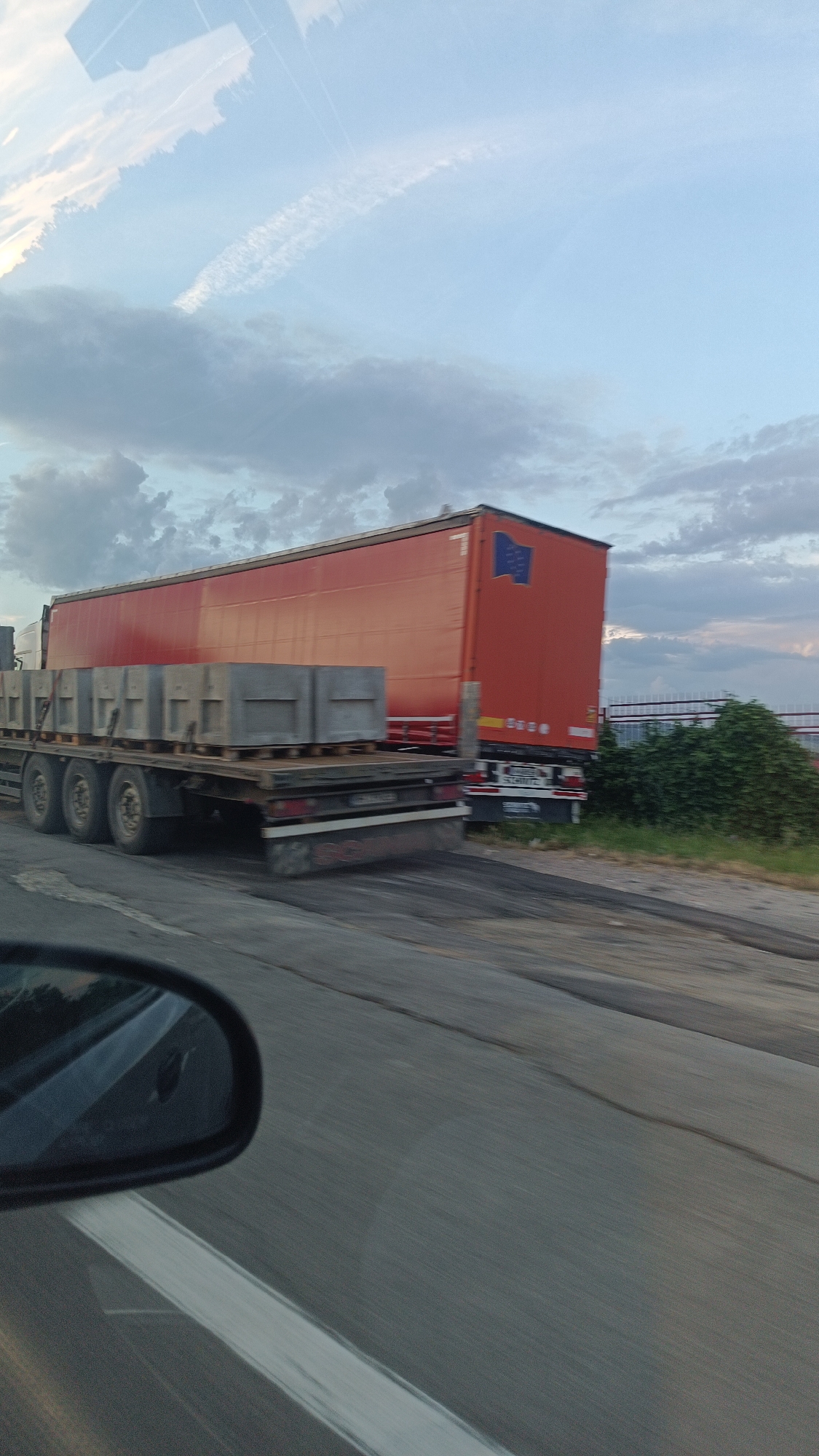 A flatbed truck carrying concrete blocks is on the road in Burgas, Bulgaria. The truck is traveling on a highway, as indicated by the road markings and the median. A large red trailer with an EU flag is parked to the side of the road. The sky is cloudy. The photo was taken from inside a vehicle as evidenced by the car mirror in the left corner of the frame.