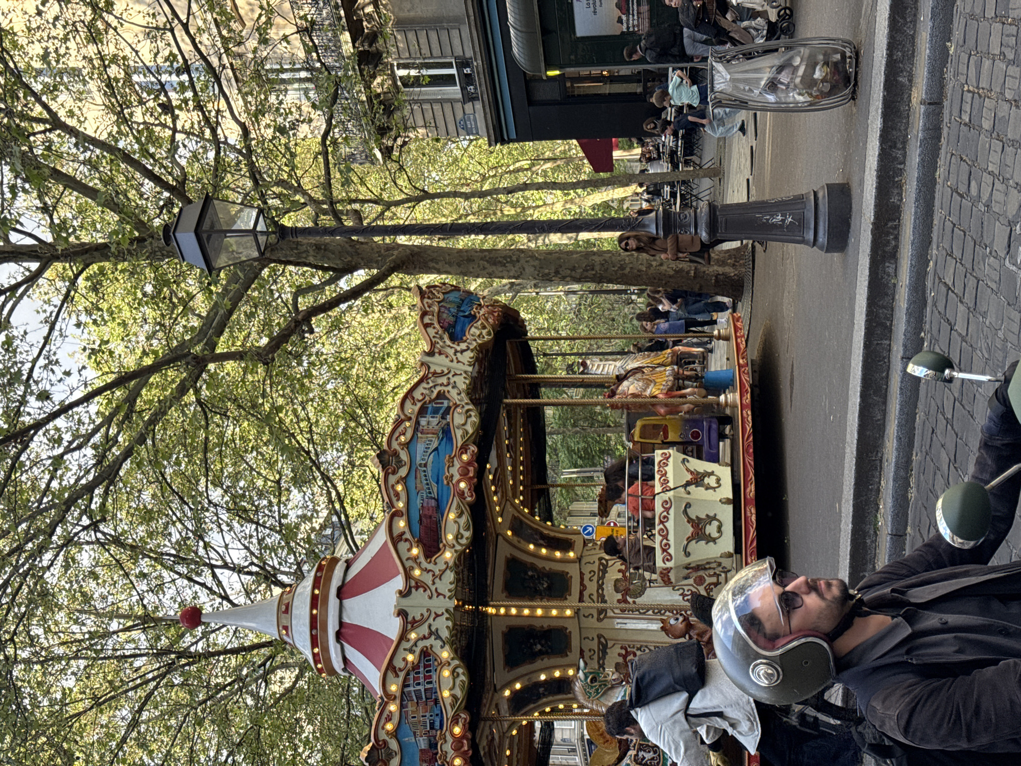 An outdoor street scene in Paris captures a lively daytime atmosphere. In the foreground, a man wearing a grey motorcycle helmet with a clear visor and sunglasses sits on a scooter or motorcycle, looking towards the right side of the frame. He has a beard and is wearing a dark jacket.
To the left of the man and occupying a significant portion of the image is a traditional, ornate carousel. It features multiple levels of painted panels depicting buildings, possibly Parisian landmarks, and is adorned with numerous warm-toned lights. The carousel has a distinctive red and white striped conical roof, topped with a red finial. Several people, including children, can be seen riding the carousel, some on classic carousel animals like horses.
The setting is characterized by mature trees with lush green leaves, suggesting a park or a tree-lined boulevard. A vintage-style black street lamp stands prominently, with its glass lantern visible among the tree branches. The ground features cobblestones in some areas and asphalt for the roadway.
In the mid-ground and background, there are classic European buildings, some with windows and dark storefronts, suggesting cafes or shops. People are visible seated at outdoor tables, enjoying the pleasant weather. A transparent public rubbish bin is also seen near a street lamp base.
Notable details include the bright daylight, indicating good weather. The overall mood is relaxed and leisurely.
Visible text includes a partially obscured sign on a building in the background that seems to read "La France révolutio..." or similar, hinting at a historical context. A small blue arrow sign, likely a traffic indicator, is visible on the carousel itself, pointing to the right. Faint white markings or graffiti are visible on the base of the street lamp.