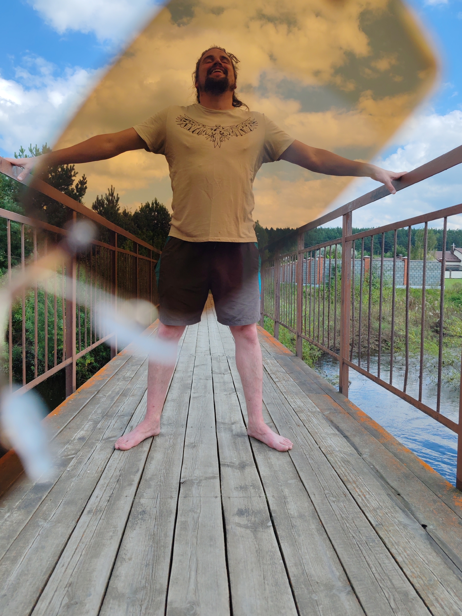 A man stands on a wooden bridge with his arms outstretched, looking up with his eyes closed and a smile on his face. He is wearing a light-colored t-shirt with a graphic design and dark shorts. The bridge has rusty metal railings and is made of weathered wooden planks. A body of water flows beneath the bridge, lined with green foliage and trees on the left. On the right side of the bridge, there is a fence, some buildings, and a cloudy sky. The upper portion of the image appears to have a filter applied, showing a diagonal split between a bright blue sky with white clouds and an orange-tinted sky with clouds. The location is identified as the city of Kashino, Russia.