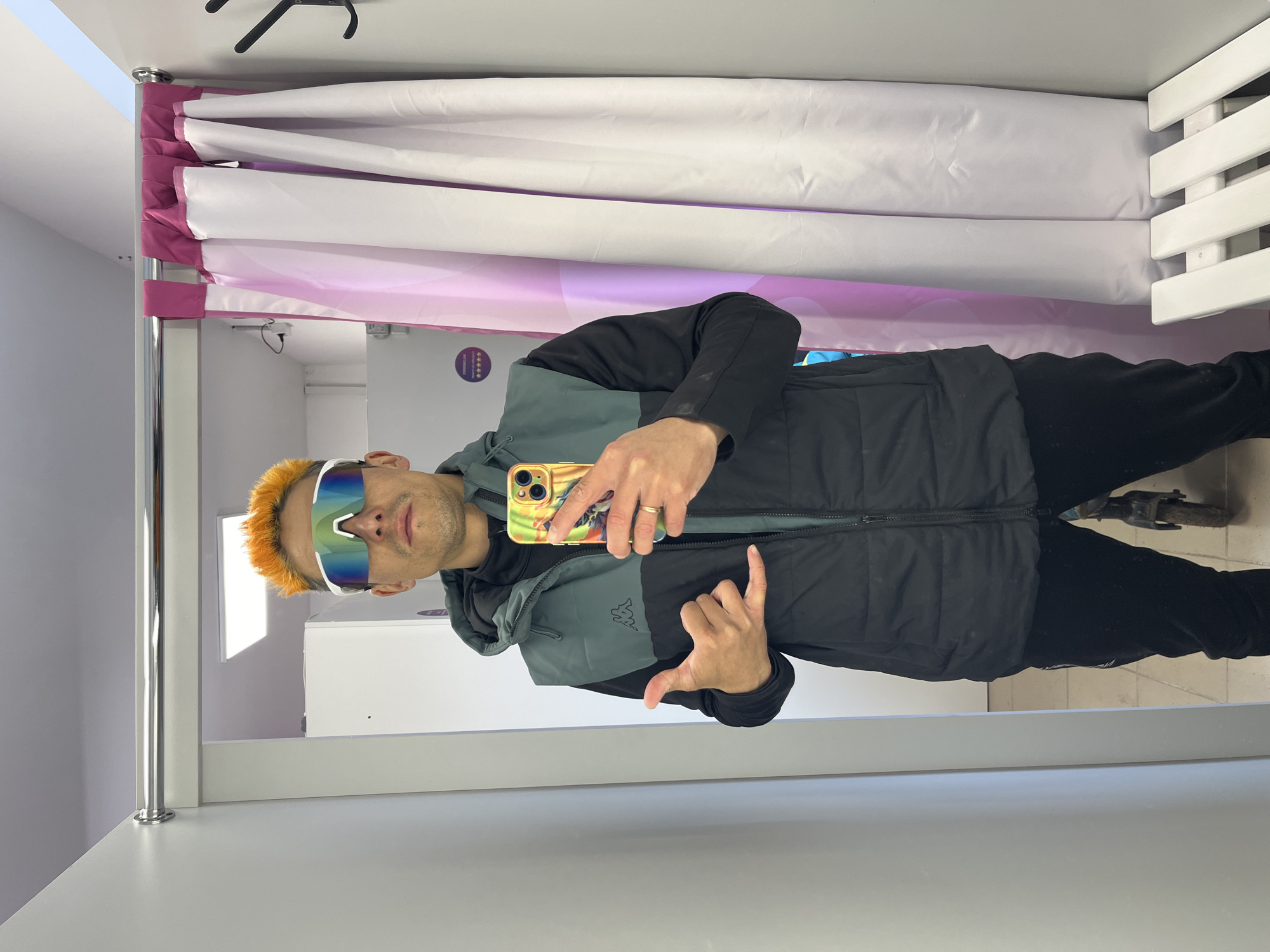A person with bright orange hair and reflective sunglasses is taking a selfie in a dressing room. They are wearing a black and gray Kappa jacket and black pants. The dressing room has white and pink curtains, white walls, and a mirror. The person is holding a gold-colored phone with a decorative case. The location is a store or boutique in Chaikovsky, Russia.