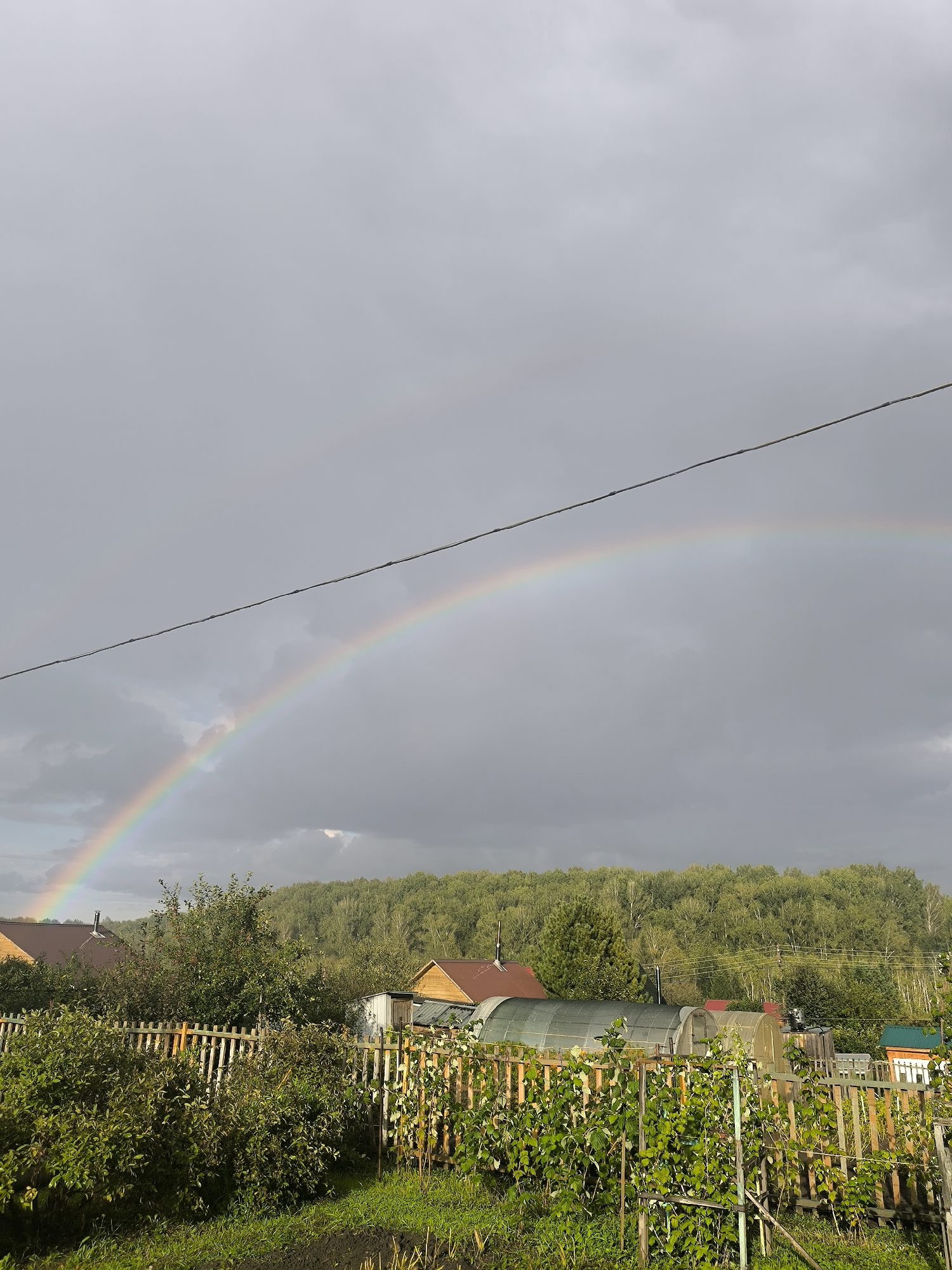 The image depicts a rural scene in the city of Maiskii, Russia, under a cloudy sky. A vibrant double rainbow is visible arching across the sky. In the foreground, a wooden fence encloses a garden with lush green vegetation. Beyond the fence, several structures are present, including houses with red roofs, greenhouses, and other small buildings. A dense forest borders the settlement in the background. A single electrical wire runs horizontally across the upper portion of the frame, intersecting the view of the rainbow. The lighting suggests it may be late afternoon or early evening, with shafts of sunlight breaking through the clouds in some areas. No people are visible in the scene.