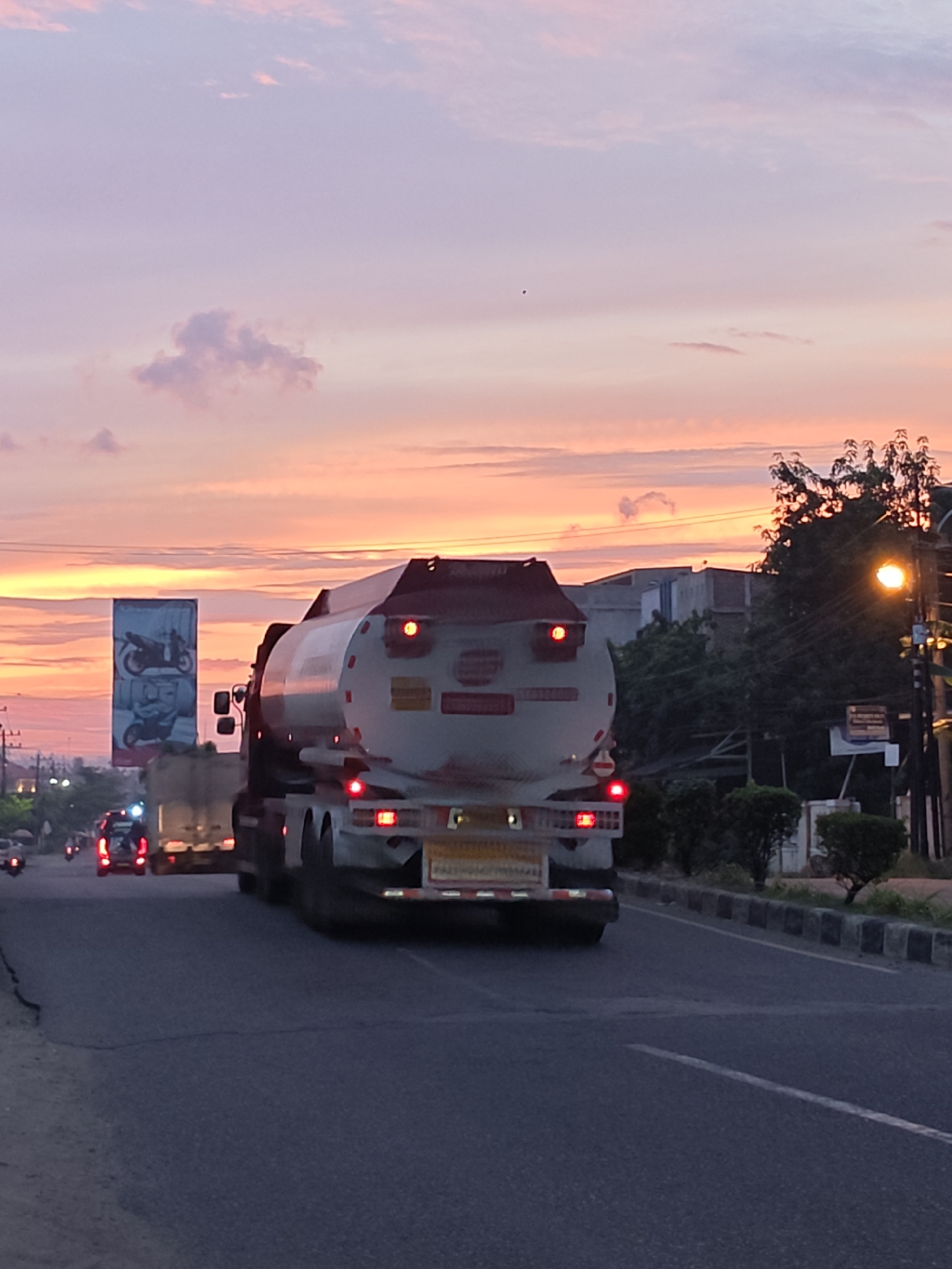 This is a medium-shot, eye-level, outdoor, vertical photograph of a road in Lhokseumawe, Indonesia, during a sunset. The sky is a gradient of soft pinks, oranges, and purples with scattered clouds. A large white tanker truck is in the foreground, traveling away from the viewer on a multi-lane road. The truck has red taillights illuminated, and a yellow sign with text is visible on its rear. To the left of the tanker truck, another truck and a red car are also on the road. In the distance, to the left, a tall billboard displaying images of motorcycles stands against the colorful sky. Trees and buildings are visible on the right side of the road, and a streetlamp with an orange glow is also present. The road itself is dark asphalt with white lane markings.