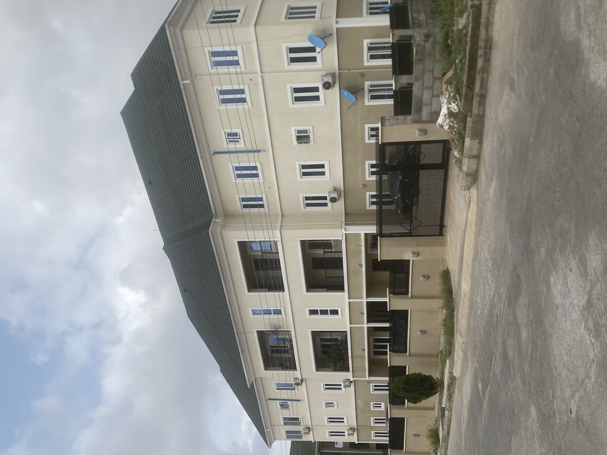 A slightly tilted eye-level shot of a multi-story residential building in Iddo Tudunwada, Nigeria, under a cloudy sky. The building is beige with white window frames and a dark green roof. Each level has balconies with black metal railings. There are air conditioning units mounted below some of the windows and blue satellite dishes attached to the upper levels. A black metal gate stands on the right side of the building, next to a small tree and some low shrubs. The foreground is a gray concrete pavement. Construction debris is piled up next to the gate.