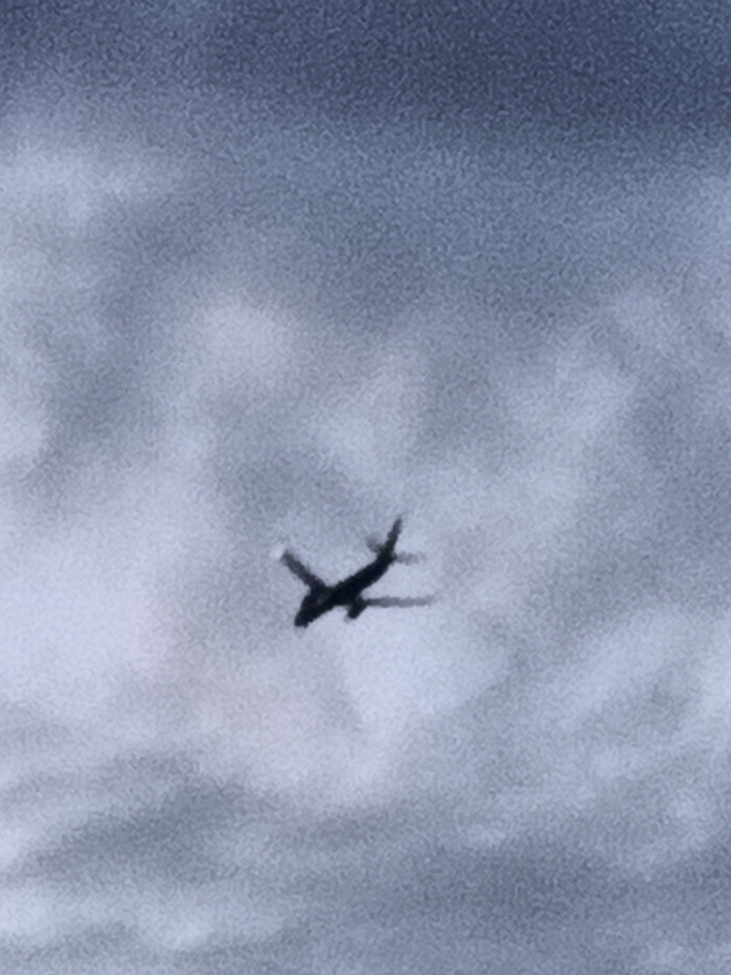 A silhouette of an airplane is captured against a cloudy sky, appearing as a dark, X-shaped form against a backdrop of muted blues and grays. The image is grainy and lacks sharp detail, making it difficult to discern specific features of the aircraft or the clouds. There are no discernible landmarks or contextual elements to indicate a specific location within Prague, Czechia. The image is an abstract representation of an airplane in flight within an overcast atmosphere.