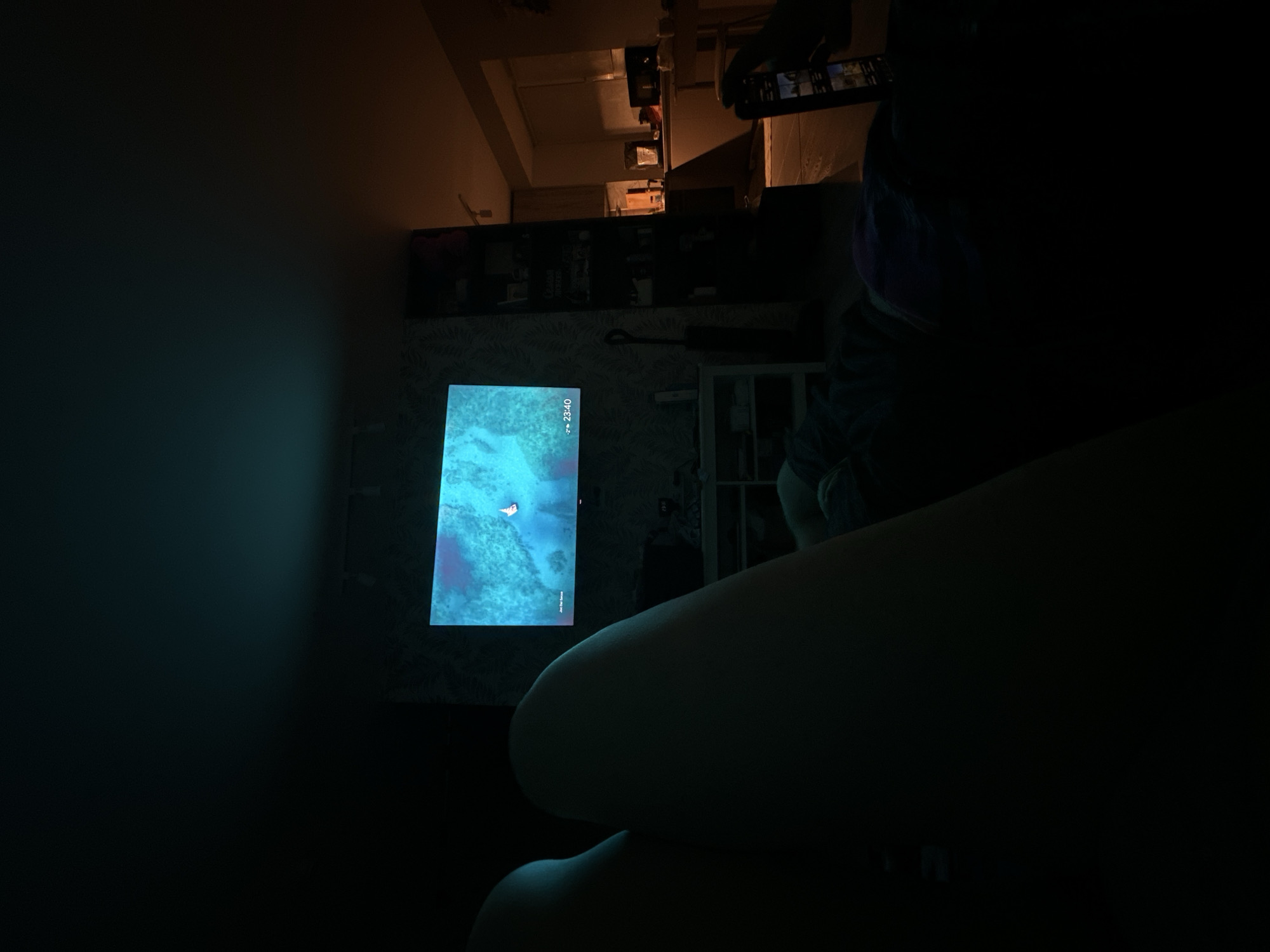 A dimly lit interior shot featuring a person seated with a glowing screen in the background displaying what appears to be a map or graphical interface. The time displayed on the screen is 23:40. Above the screen, there is wallpaper and what might be a shelf or bookcase with miscellaneous items. At the top of the frame, an inverted view into another room is visible, containing furniture and objects, presumably taken by a phone. The person holding the phone is also in the foreground. I cannot determine the location to be Chaikovsky, Russia, with the given information.