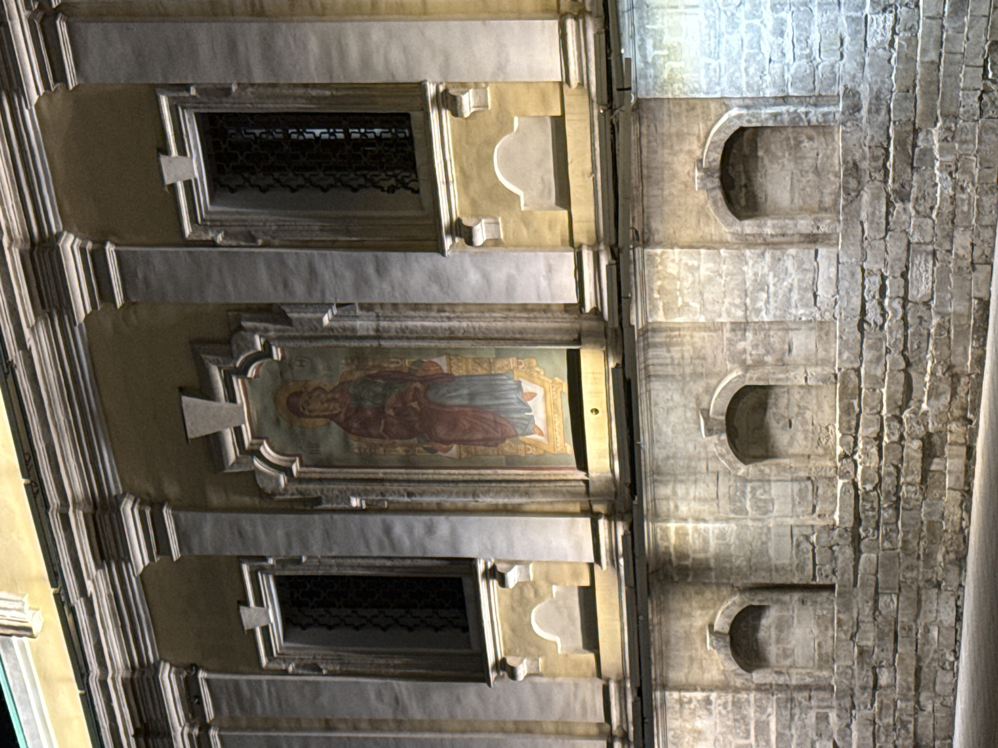 The image depicts the facade of a building at night, illuminated by artificial lighting. The facade is adorned with architectural elements such as columns, cornices, and decorative niches. There are three prominent rectangular windows, two on the sides and one central. The central window contains a fresco or mural depicting a figure in religious attire. The lower part of the building features a wall constructed of rough-hewn stones, with three arched alcoves inset into it. The building appears to be old, with a weathered appearance. The overall style suggests a historical religious or public building. This facade is located at the St. Andrew's Cathedral in Moscow, Russia.